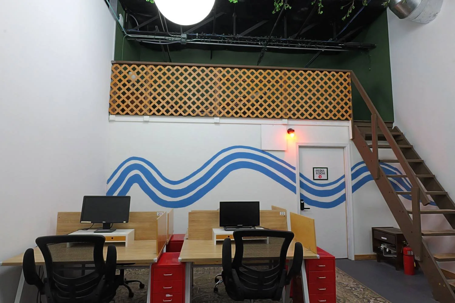 Office workspace with two desks separated by dividers, each with a computer monitor and office chair, against a wall with blue wave mural, a staircase to a loft area, and a media room door.
