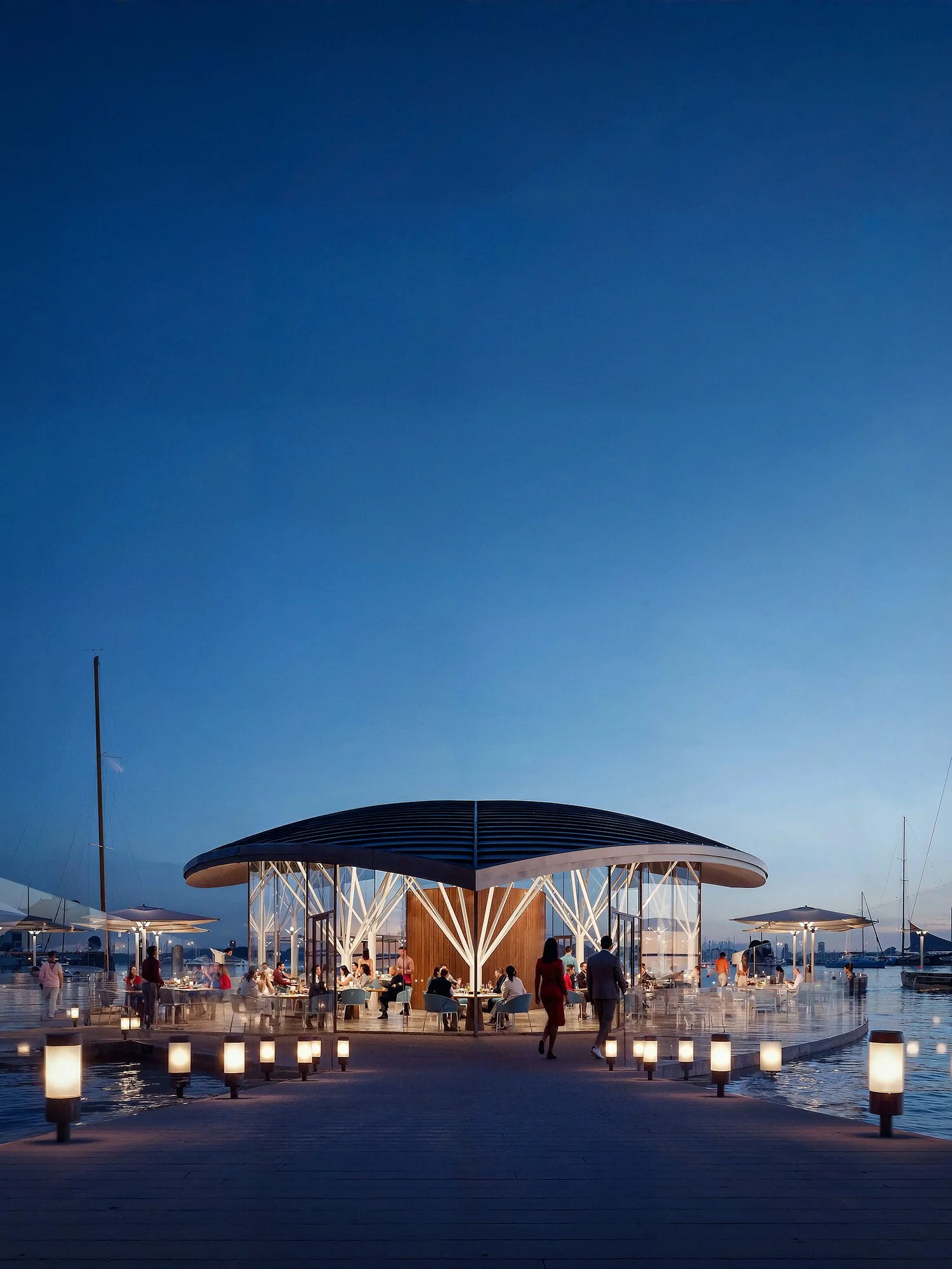 An outdoor waterfront dining area during evening with people seated, illuminated by lanterns, with a modern pavilion structure and sailboats docked in the background under a darkening sky.