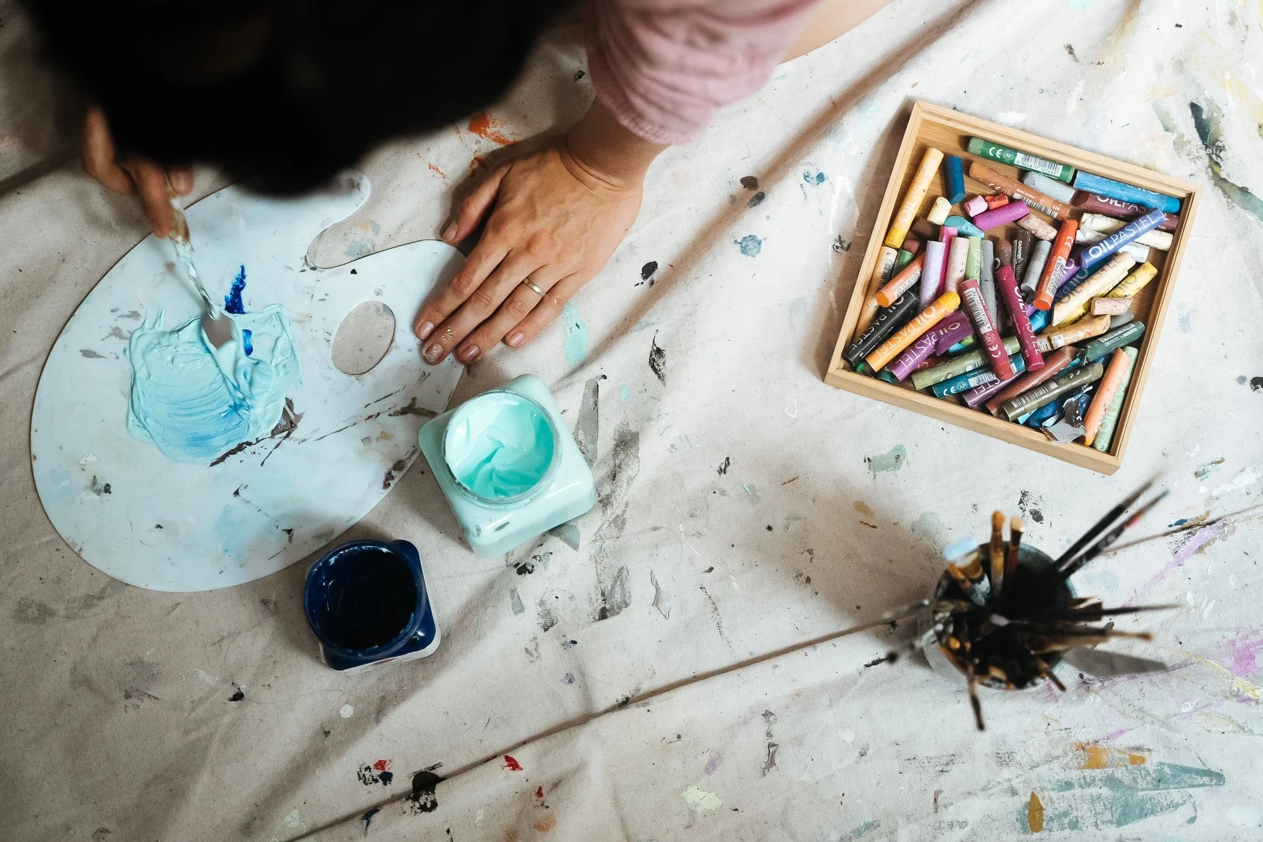 Person creating art with blue paint, with a box of colorful chalks nearby on a messy table.