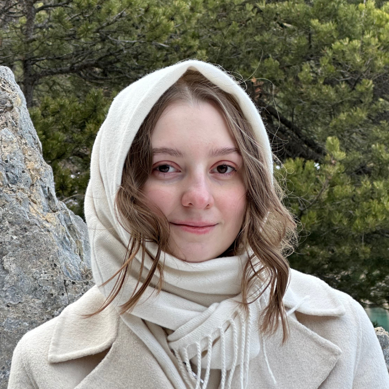 Marie Cela, a white person wearing a light coat, scarf, and hood outdoors.