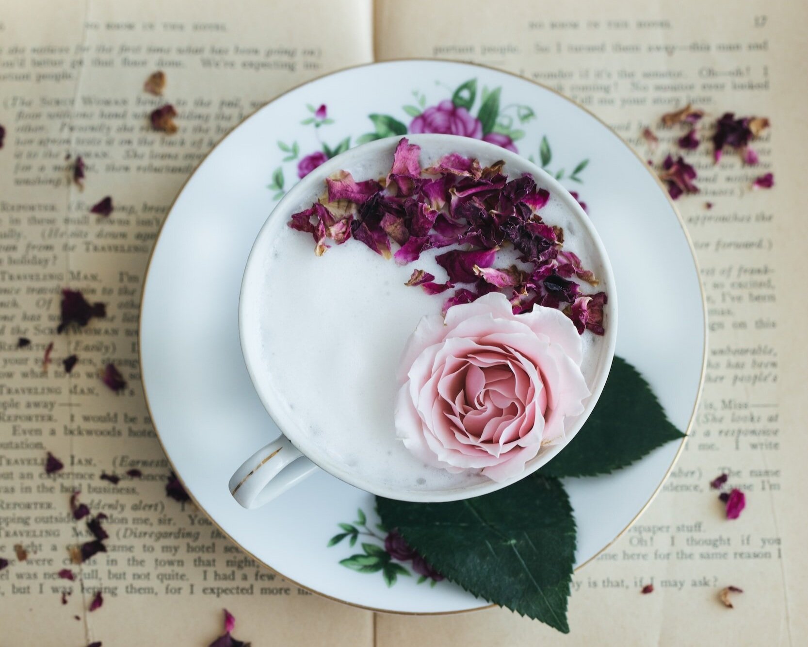 Overhead view of an open book, on top of which sits a white cup and saucer; dark and light-pink flowers float on the foam inside.