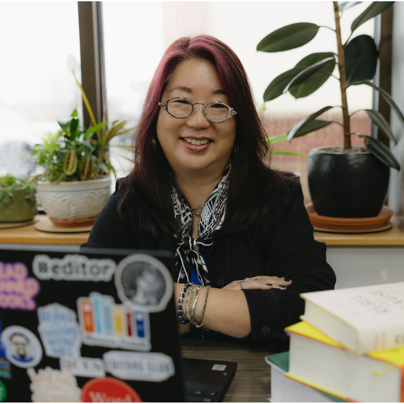 Erica Mito, an Asian person with glasses smiling at a desk beside books and a laptop.