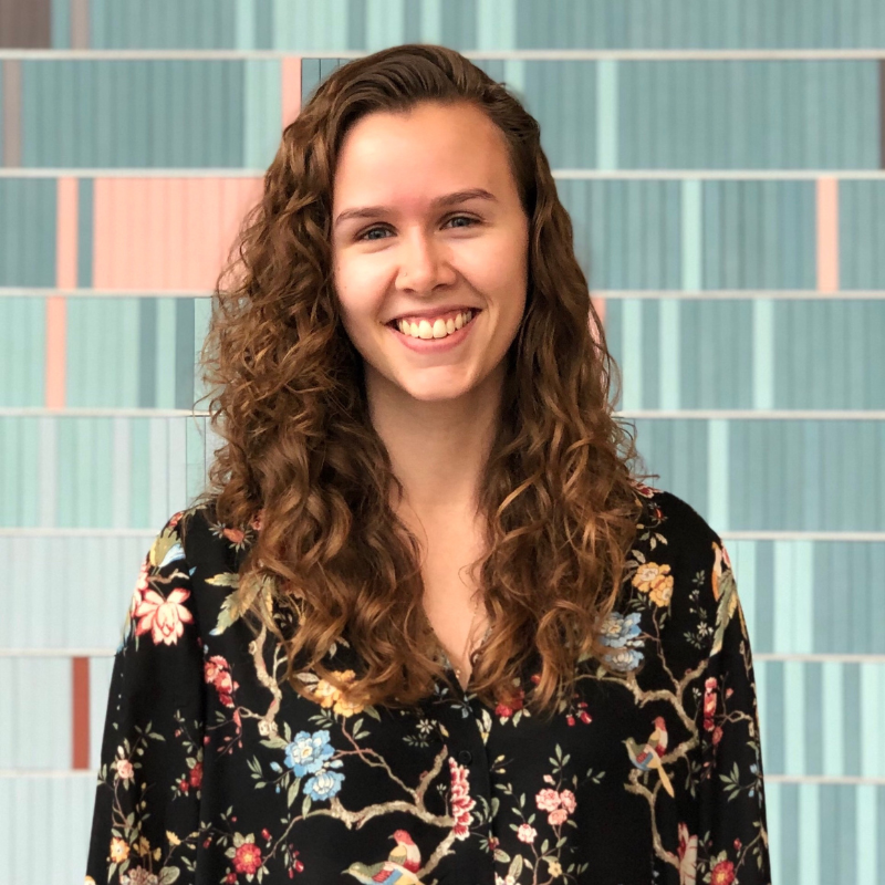 Brenna Bailey-Davies, a white person with long curly brown hair smiling.