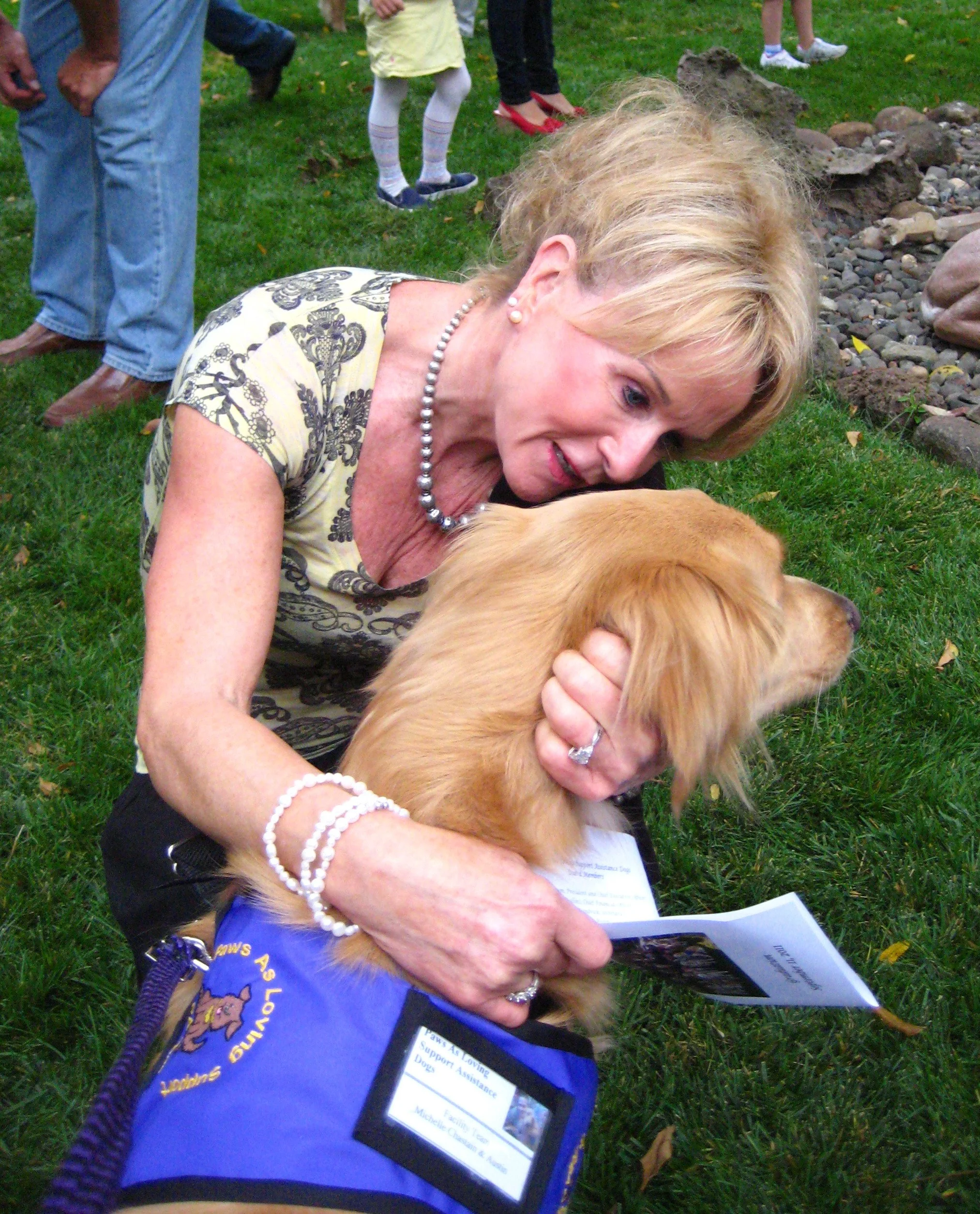 A woman with blonde hair, pearl earrings, and a pearl necklace is hugging a golden retriever dog while reading a document outdoors on grass. Other people are standing nearby in the background.