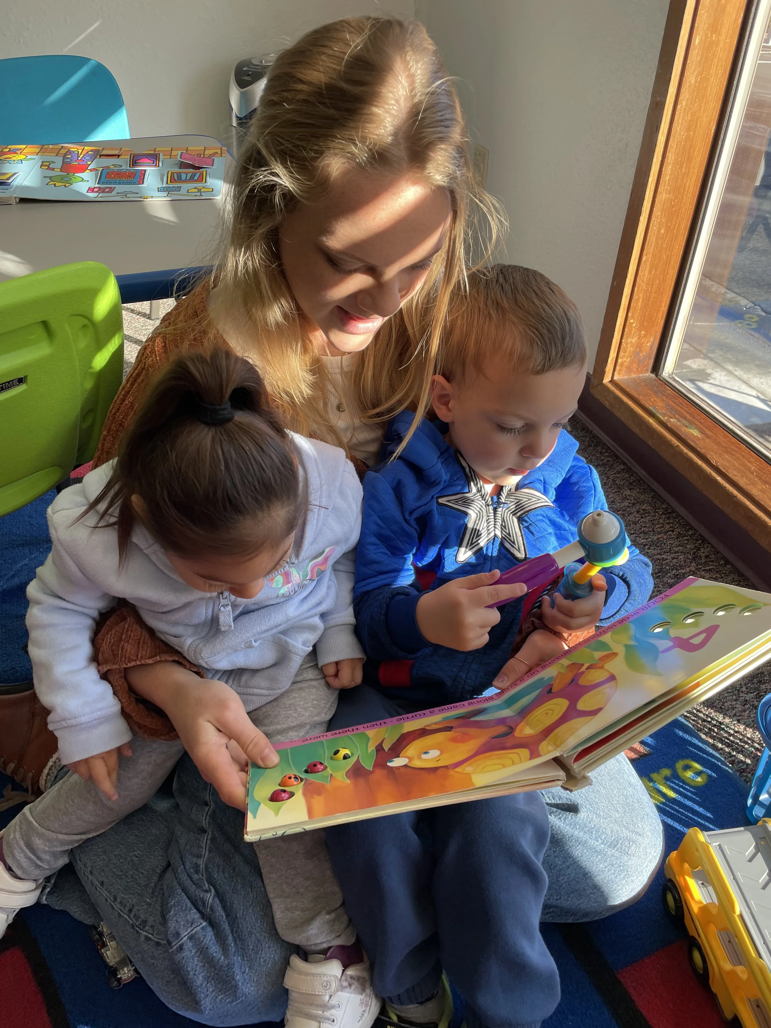 A woman with blond hair and two children sitting together and looking at a large colorful children's book with cartoon characters. The girl in front has brown hair tied in a ponytail, and the boy has light brown hair, wearing a blue jacket, holding a toy mic. There are toys and a window with sunlight in the background.