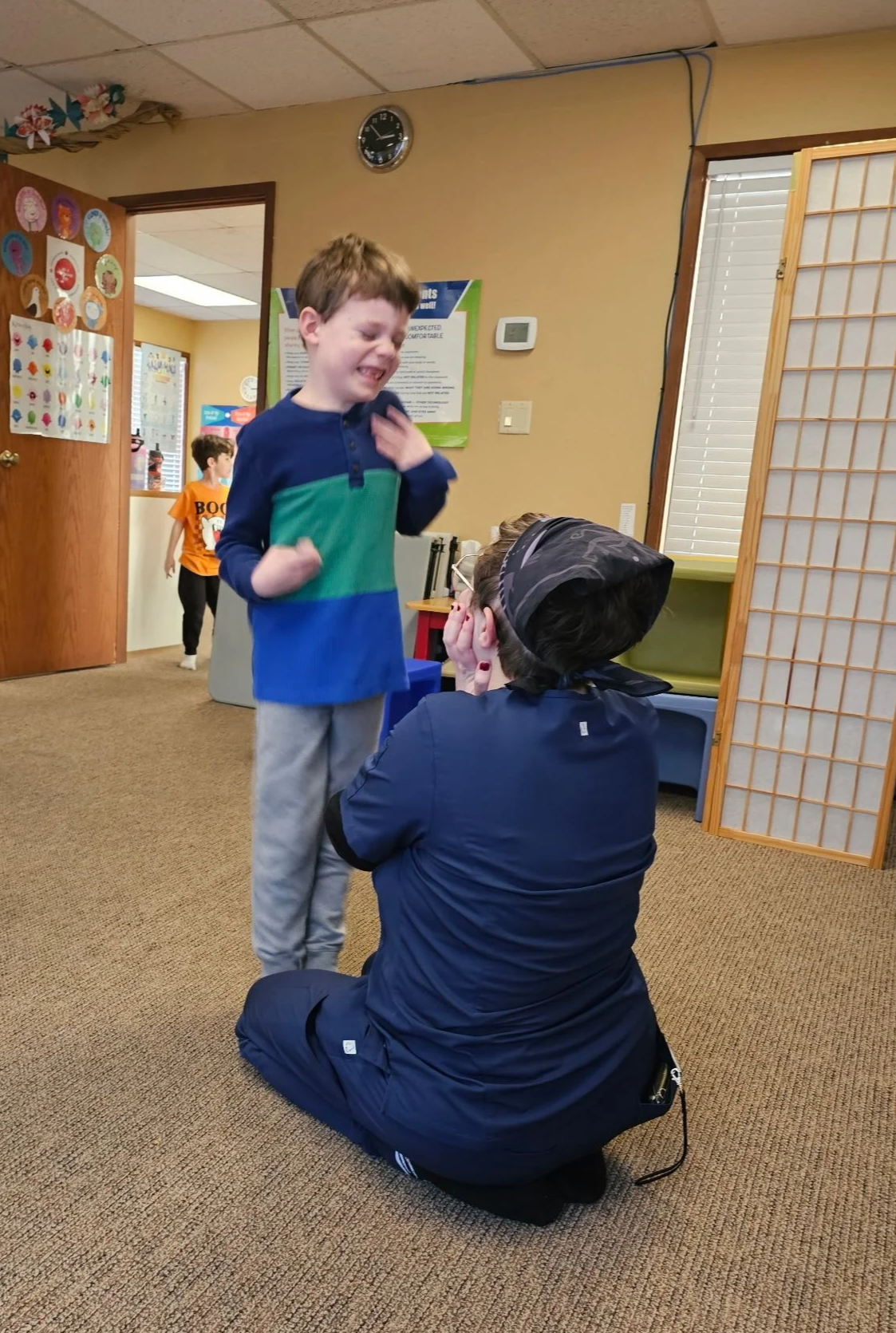 A young boy and a woman in a headscarf share an emotional moment inside a classroom with children in the background. The boy appears to be crying or upset, and the woman, kneeling on the carpeted floor, is comforting him.