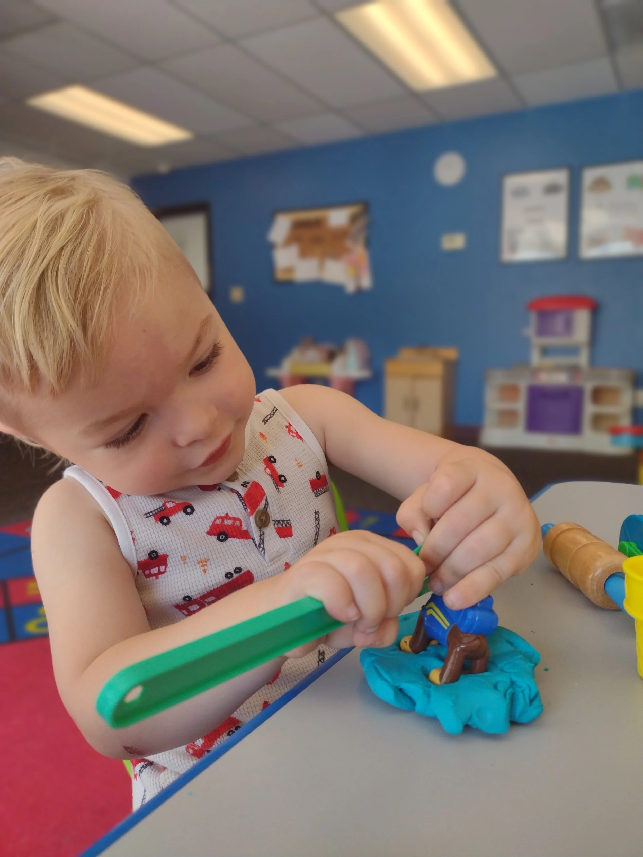A young child with blonde hair wearing a white sleeveless shirt with red and black vehicle prints, engaged in playing with colorful modeling clay on a table. The background shows a classroom with blue walls, bulletin boards, and play furniture.