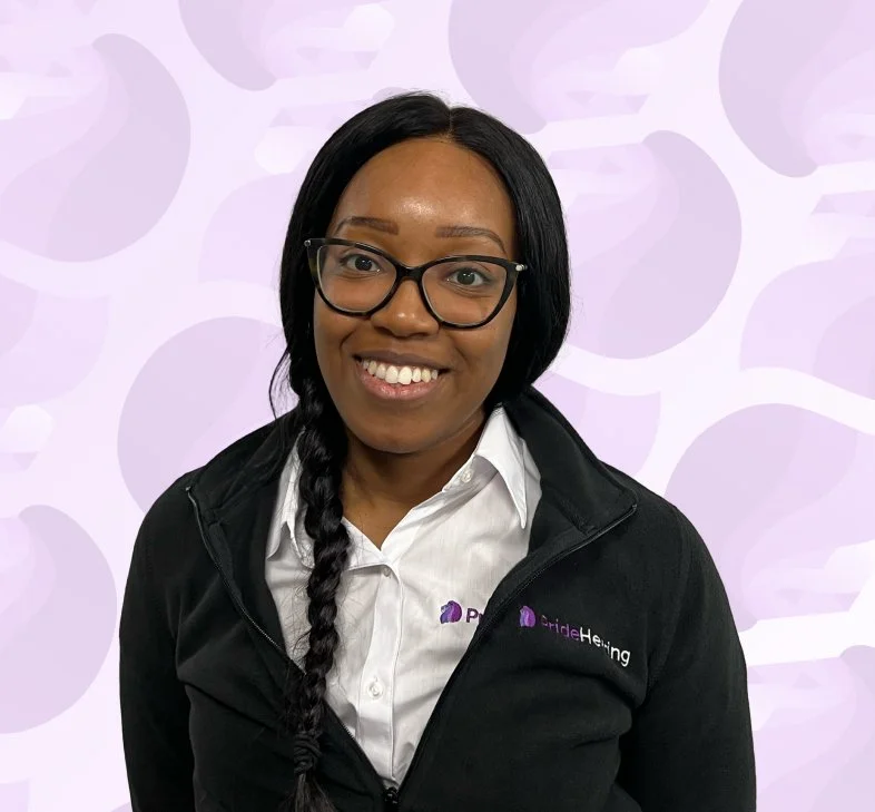 A smiling woman with black hair in a braid, wearing glasses, a white collared shirt, and a black jacket with a logo, standing against a light purple background with a pattern of circles.