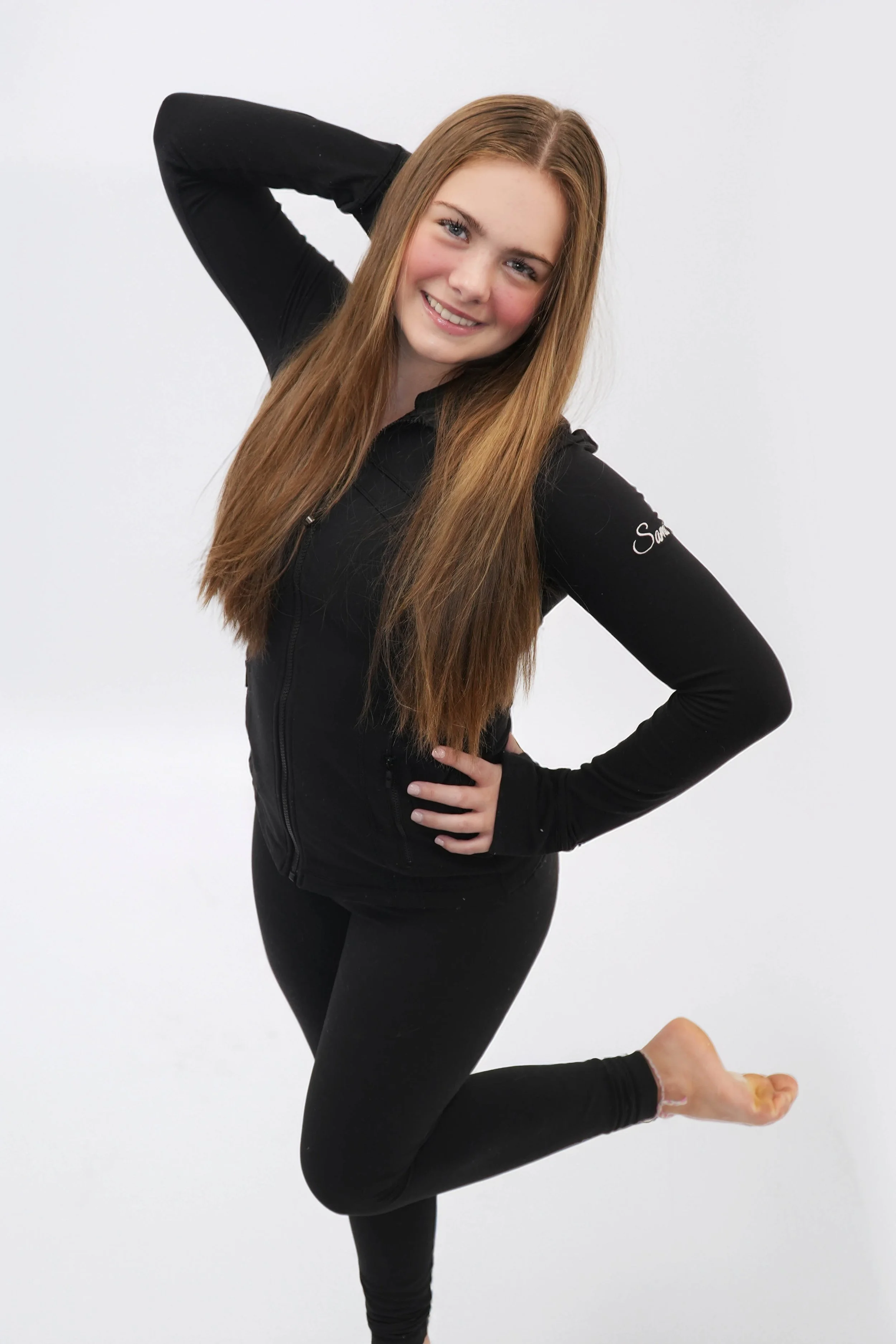 Young woman with long red hair wearing a white shirt and pants, posing with her left arm extended above her head against a dark background with green lighting.