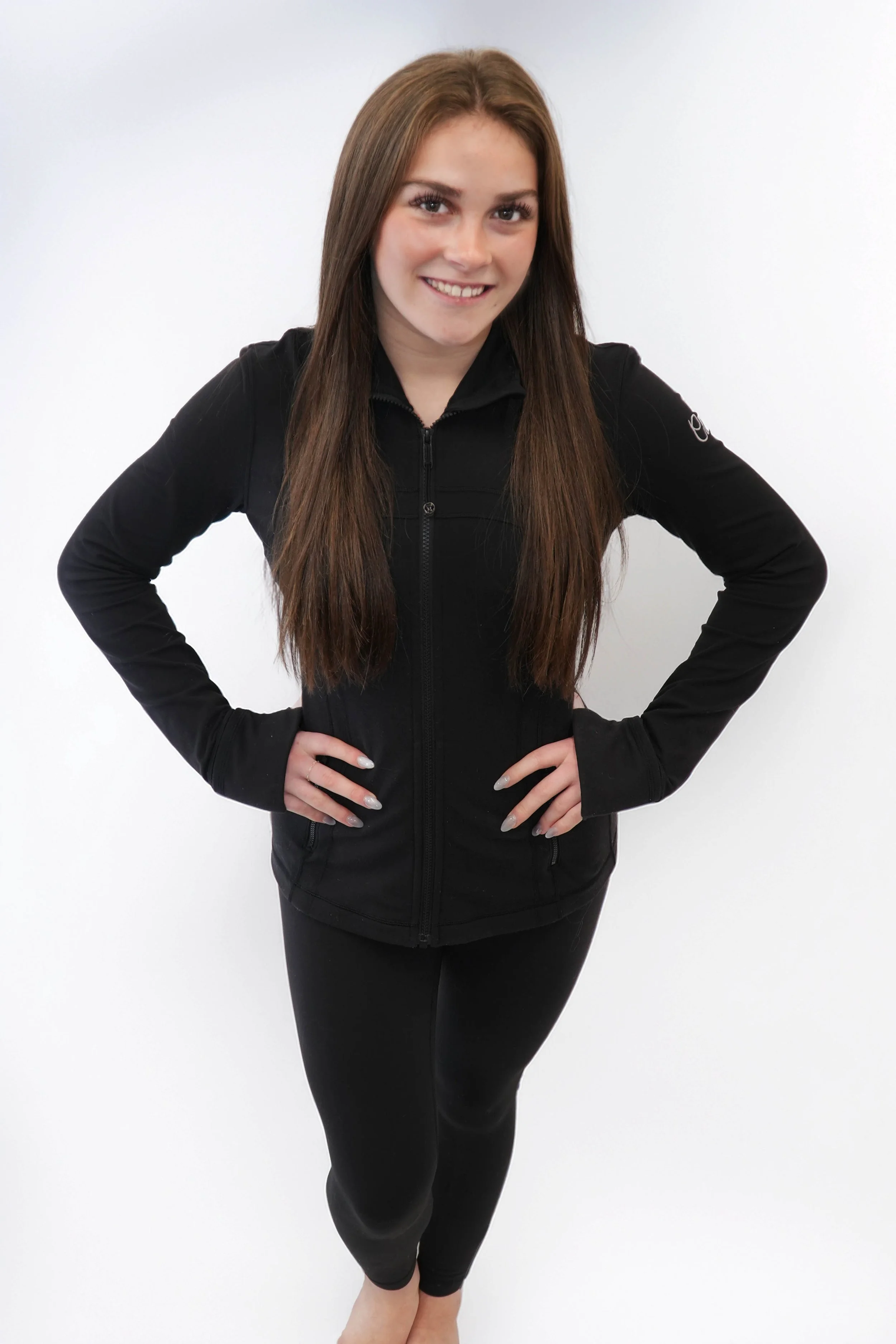 A young woman with long wavy brown hair in a white long-sleeve crop top and matching sweatpants, kneeling on a dark surface against a dark background, with green lighting illuminating her face and clothing.