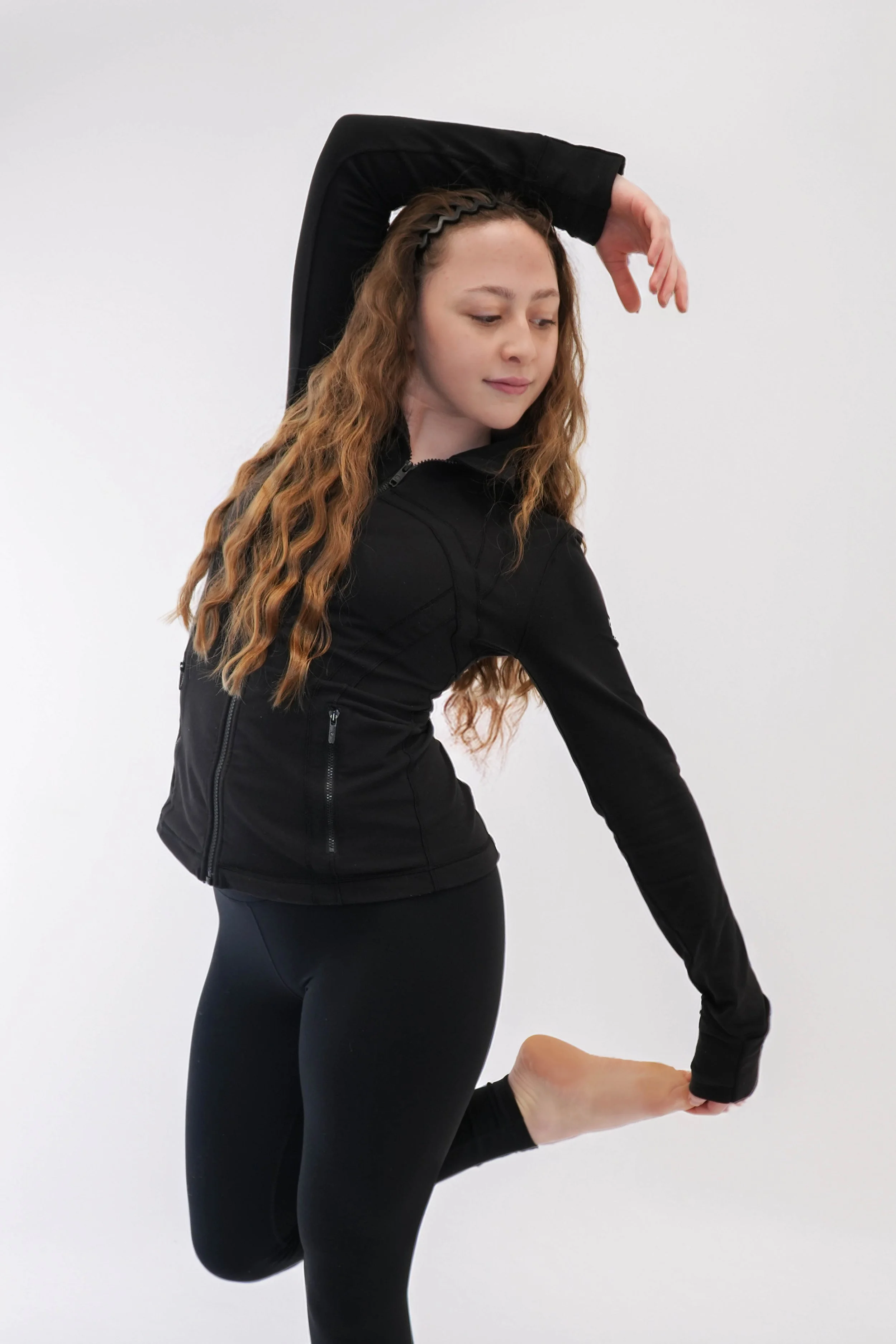 A woman with wavy hair wearing a white long-sleeve top and white pants, stretching with arms extended overhead against a dark background, illuminated by green and pink lights.