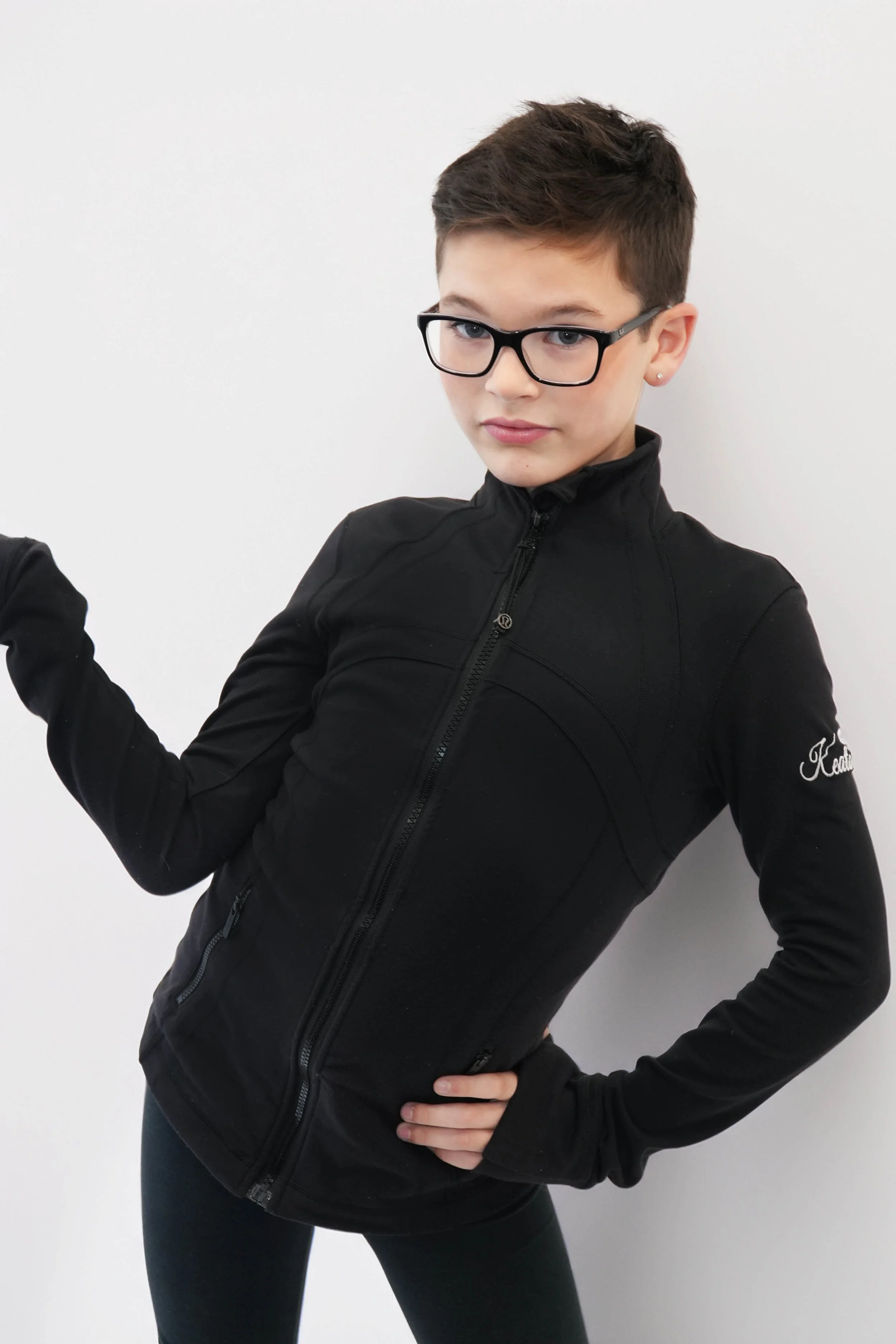 A young person with short dark hair, dressed in a white jacket and white shirt, is lying on the ground with one hand flat on the surface and the other arm extended upwards. The background is black, and the person is illuminated with green lighting.