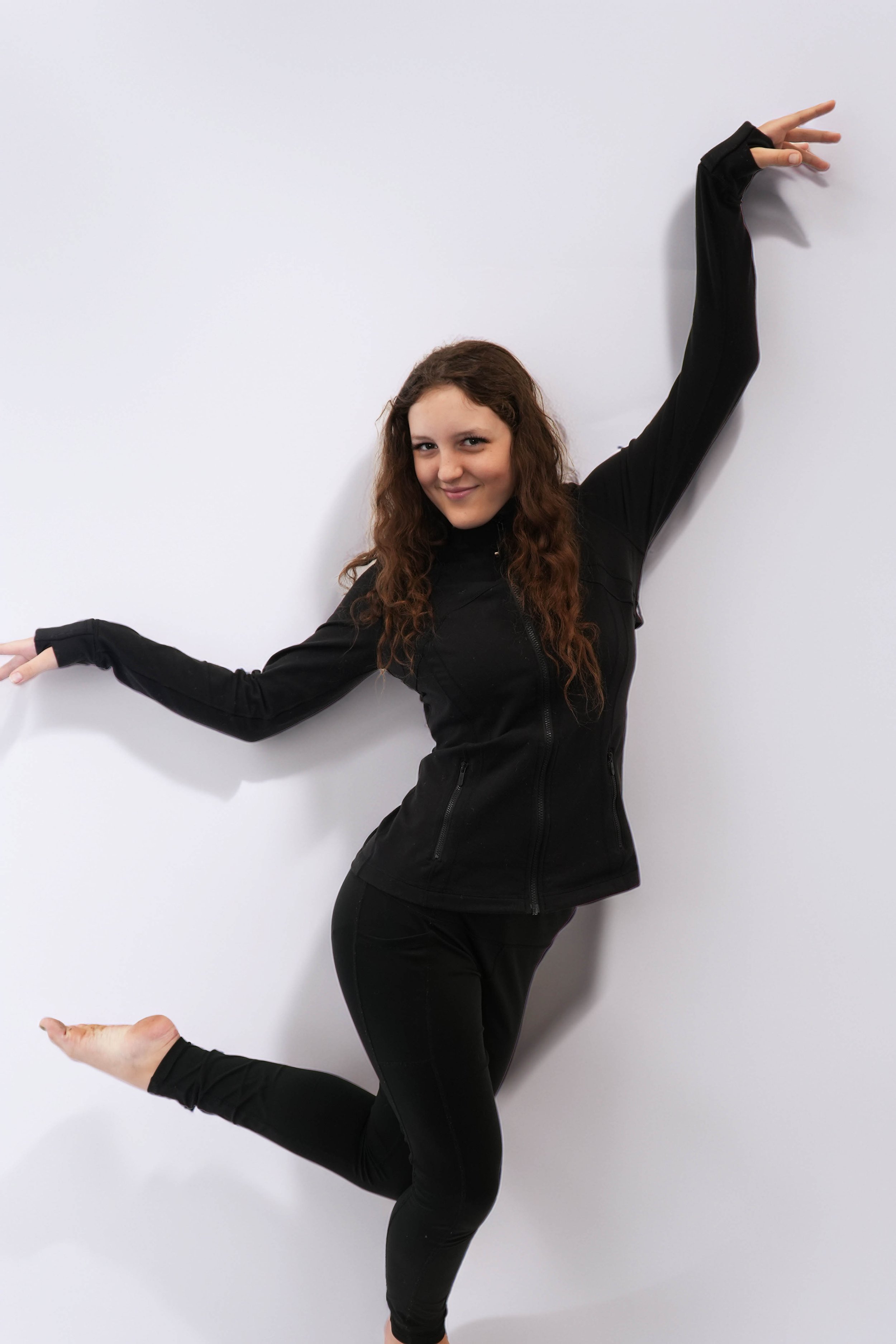 Young woman with long, curly brown hair, wearing a cream-colored long sleeve top and matching pants, posed against a black background with green lighting.