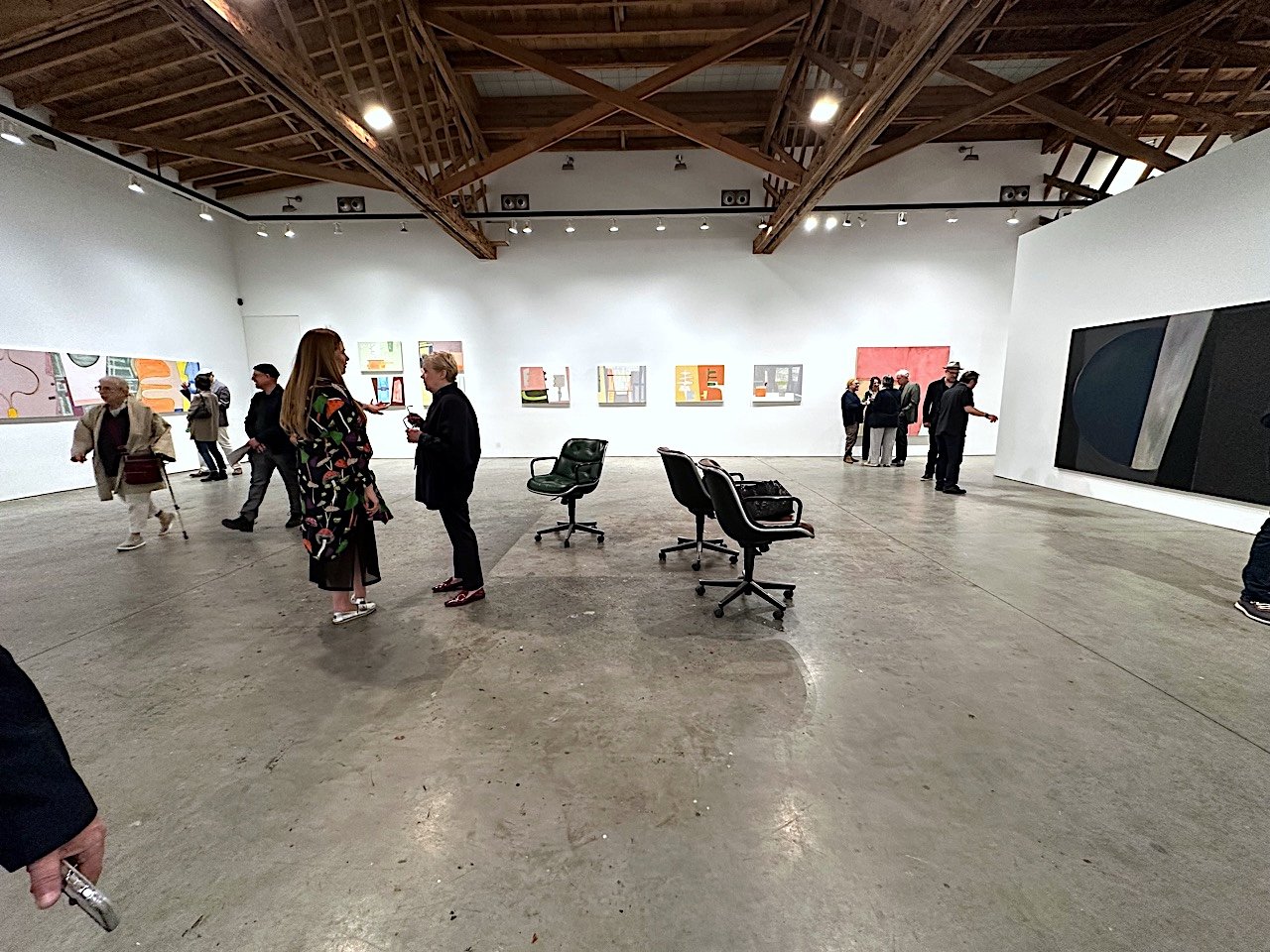 Art gallery interior with people viewing paintings on white walls, exposed wooden ceiling, and office chairs around the room.