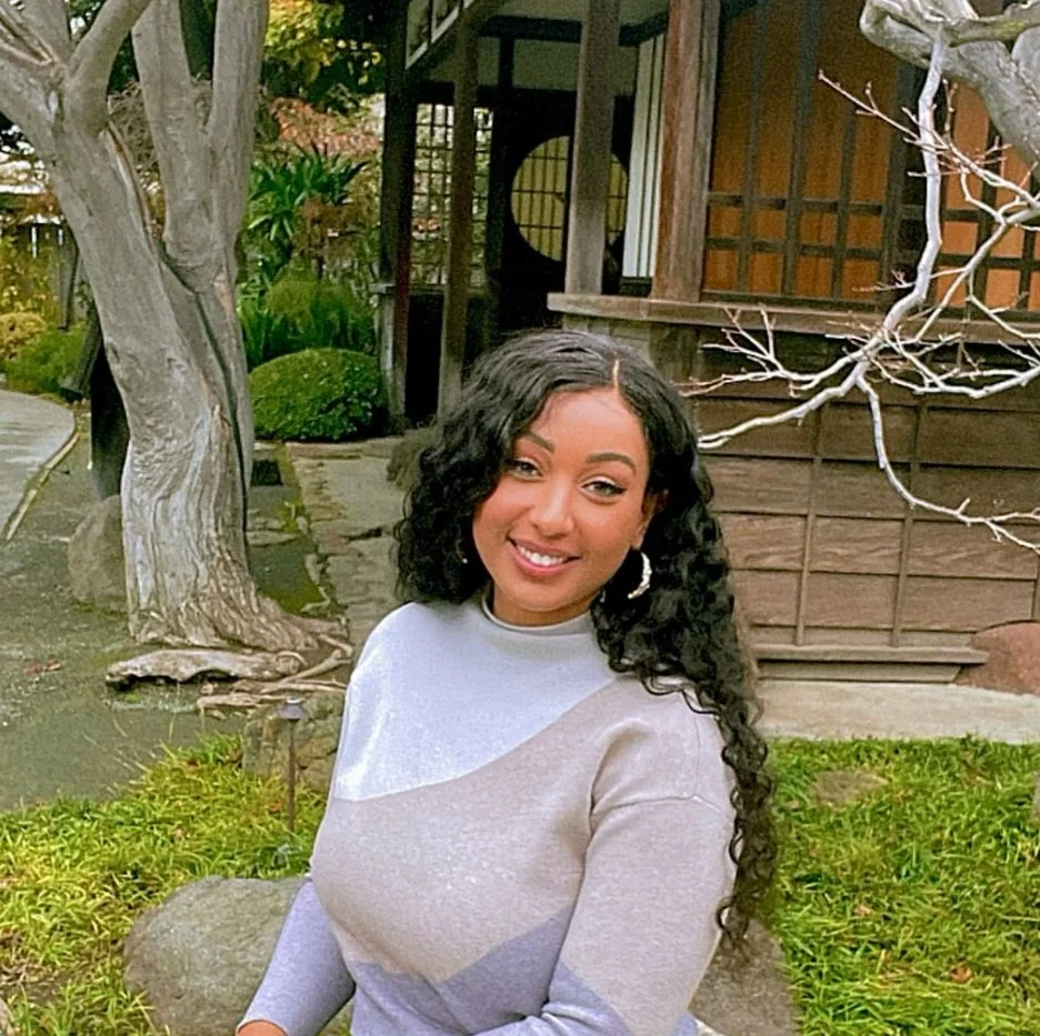 A young woman with curly black hair sitting outdoors near a tree, smiling while looking at the camera, with greenery and a wooden building in the background.