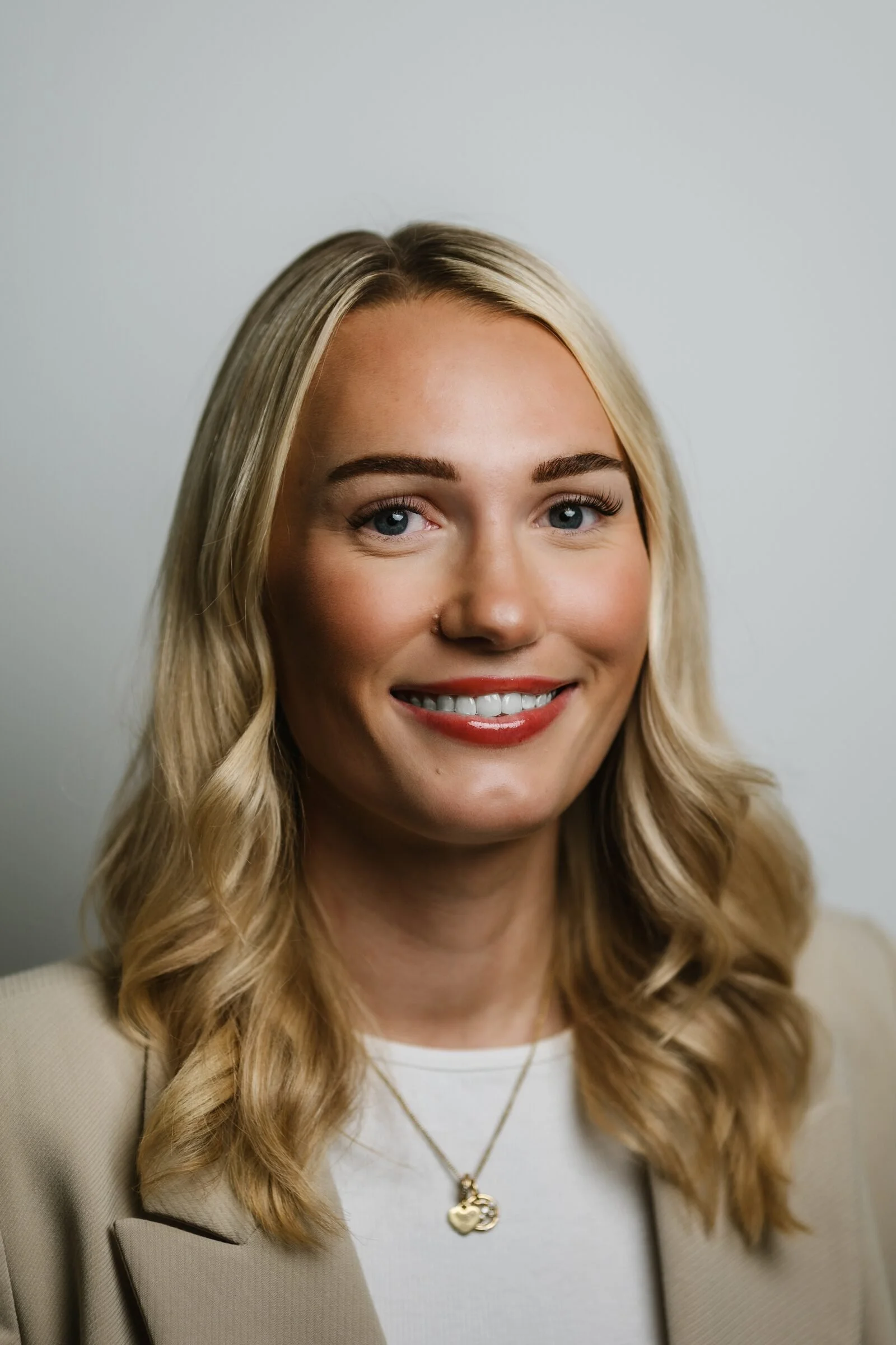 Professional headshot of a smiling woman with long blonde hair and a beige blazer against a plain light grey background.