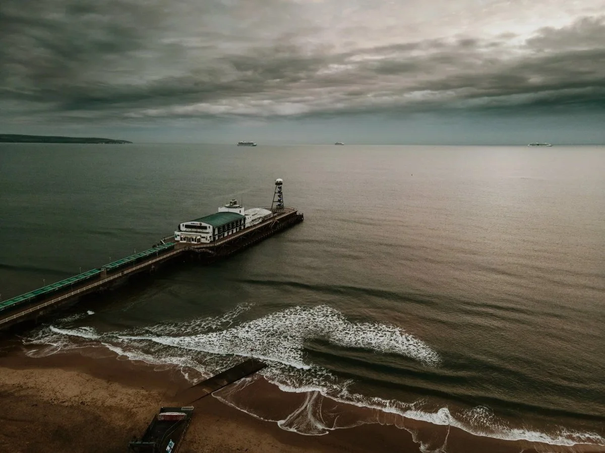 Aerial view of a quiet sandy beach and pier under a dramatic grey sky, with waves rolling into the shore