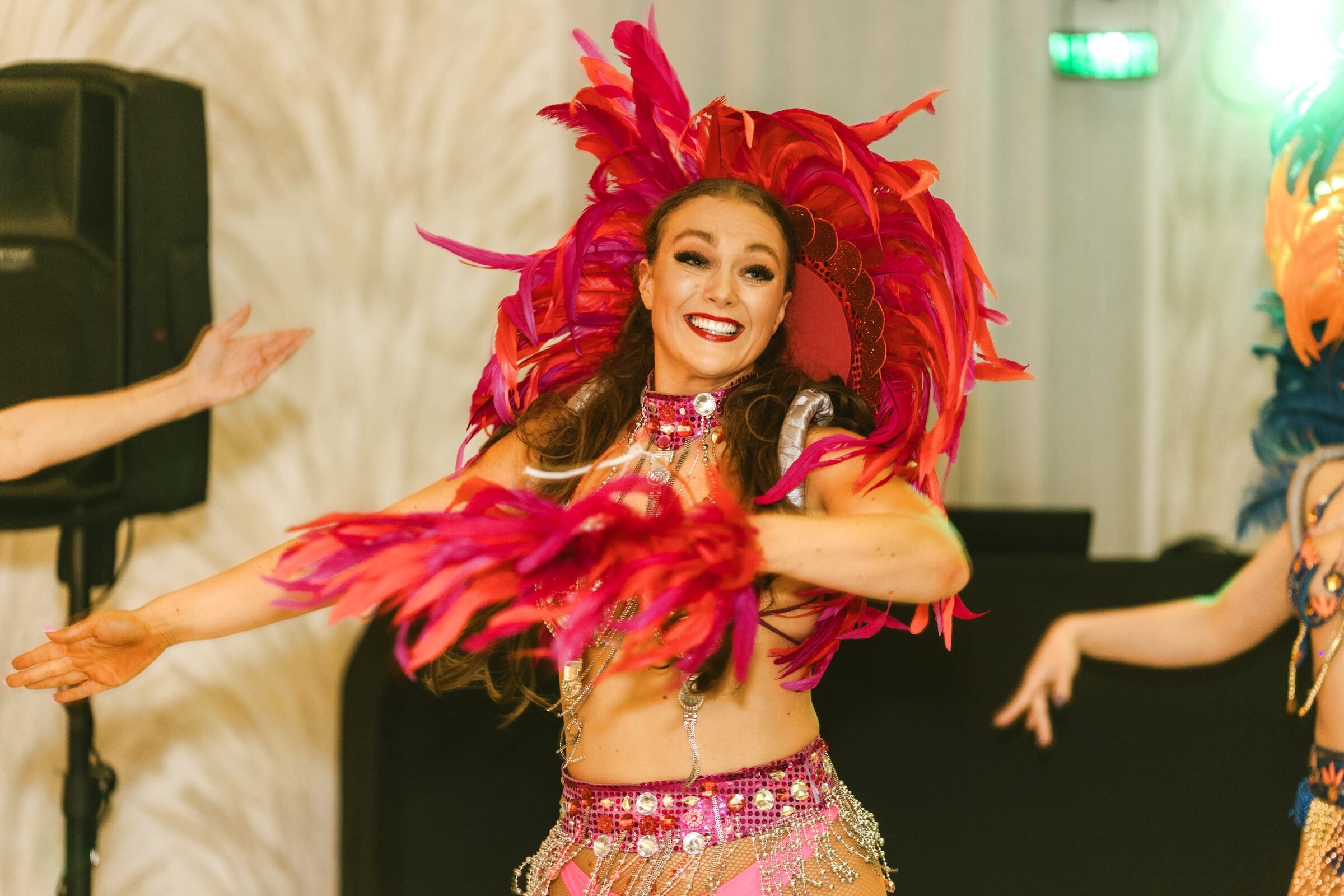 Event photo of a smiling dancer in a red feathered headdress and costume, mid-performance under stage lights.