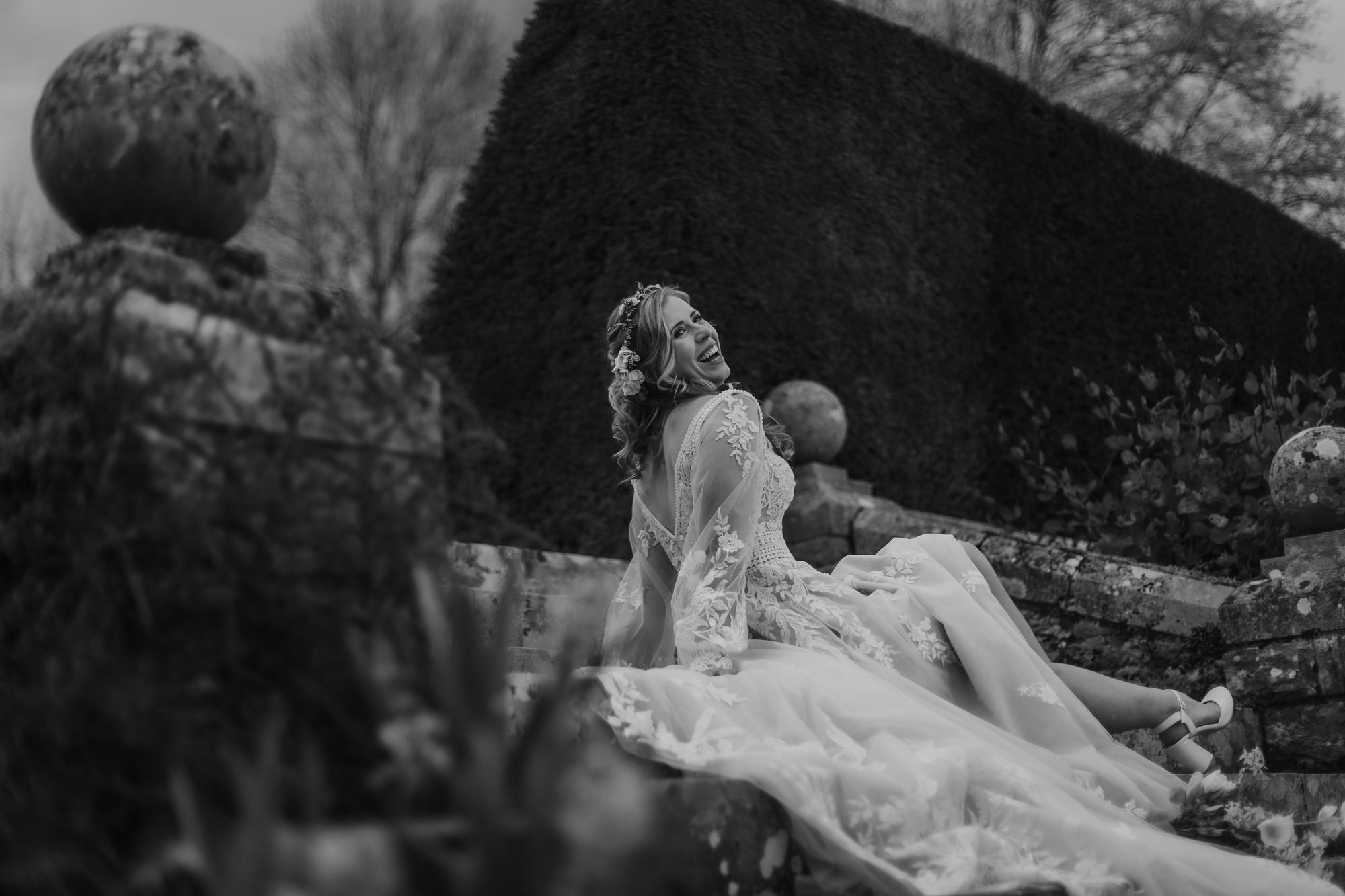 Black and white photo of a bride leaning back on a stone garden wall, laughing