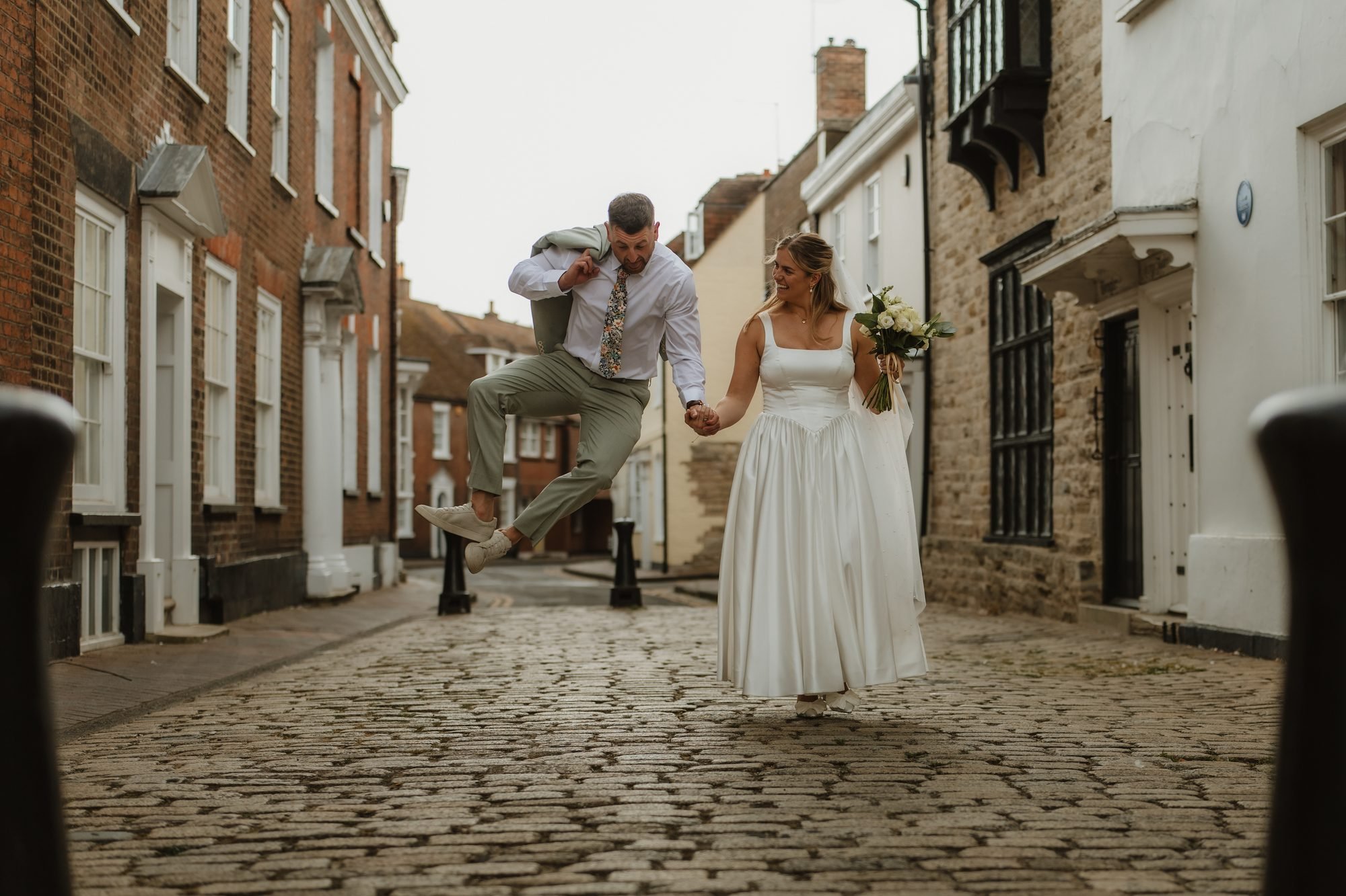 Groom jumping mid-air beside his bride on a cobbled street