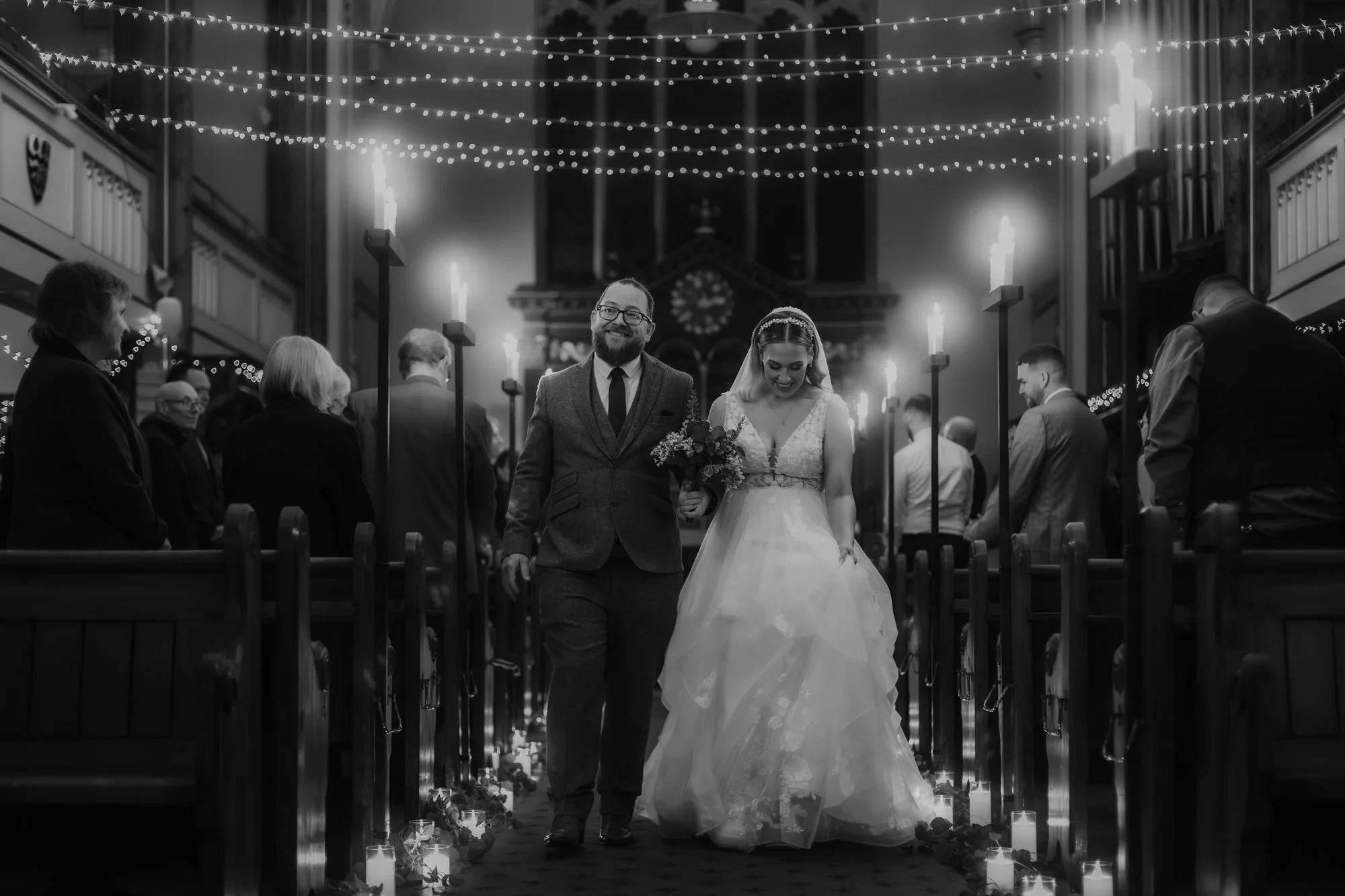 Black and white photo of a bride and groom walking down a candlelit church aisle