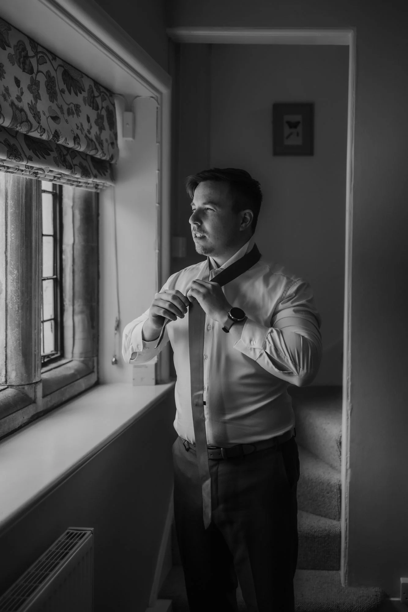 Black and white photo of a groom adjusting his collar by the window