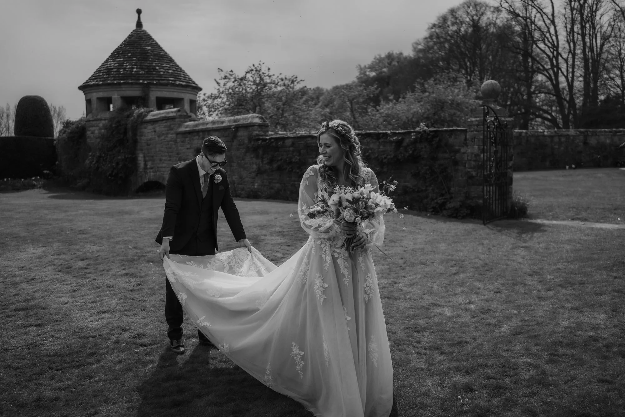 Black and white photo of a groom sweeping out the train of the bride's dress in a walled garden