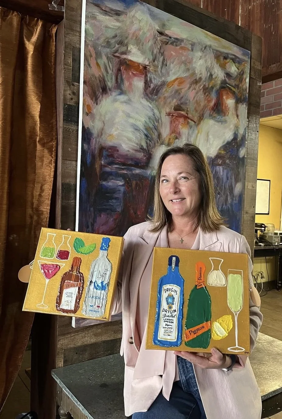 A woman holding two colorful paintings of alcohol bottles and glasses in front of a large abstract painting in a gallery setting.