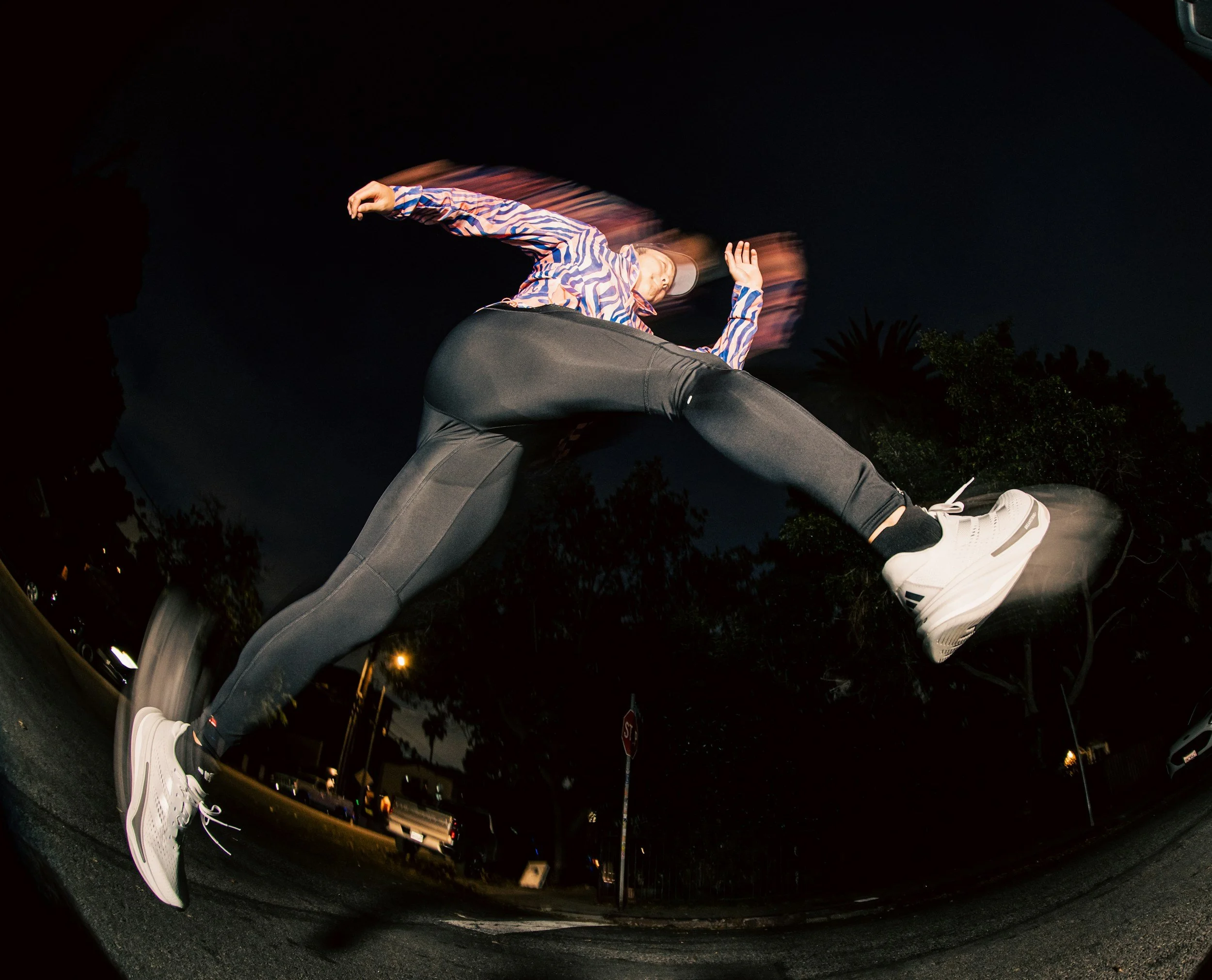 A person mid-air, captured in a dynamic jump at night, wearing a colorful striped shirt, black pants, and white sneakers, in an outdoor setting with trees and streetlights.