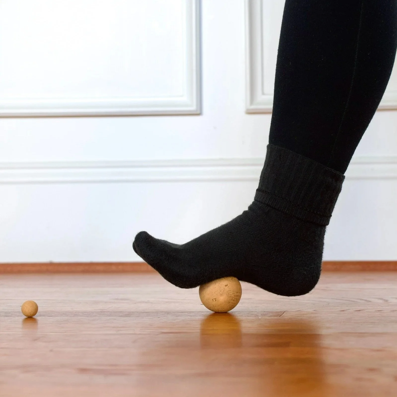 Person wearing black sock balancing on a wooden balancing ball on a hardwood floor, with a smaller wooden ball nearby.