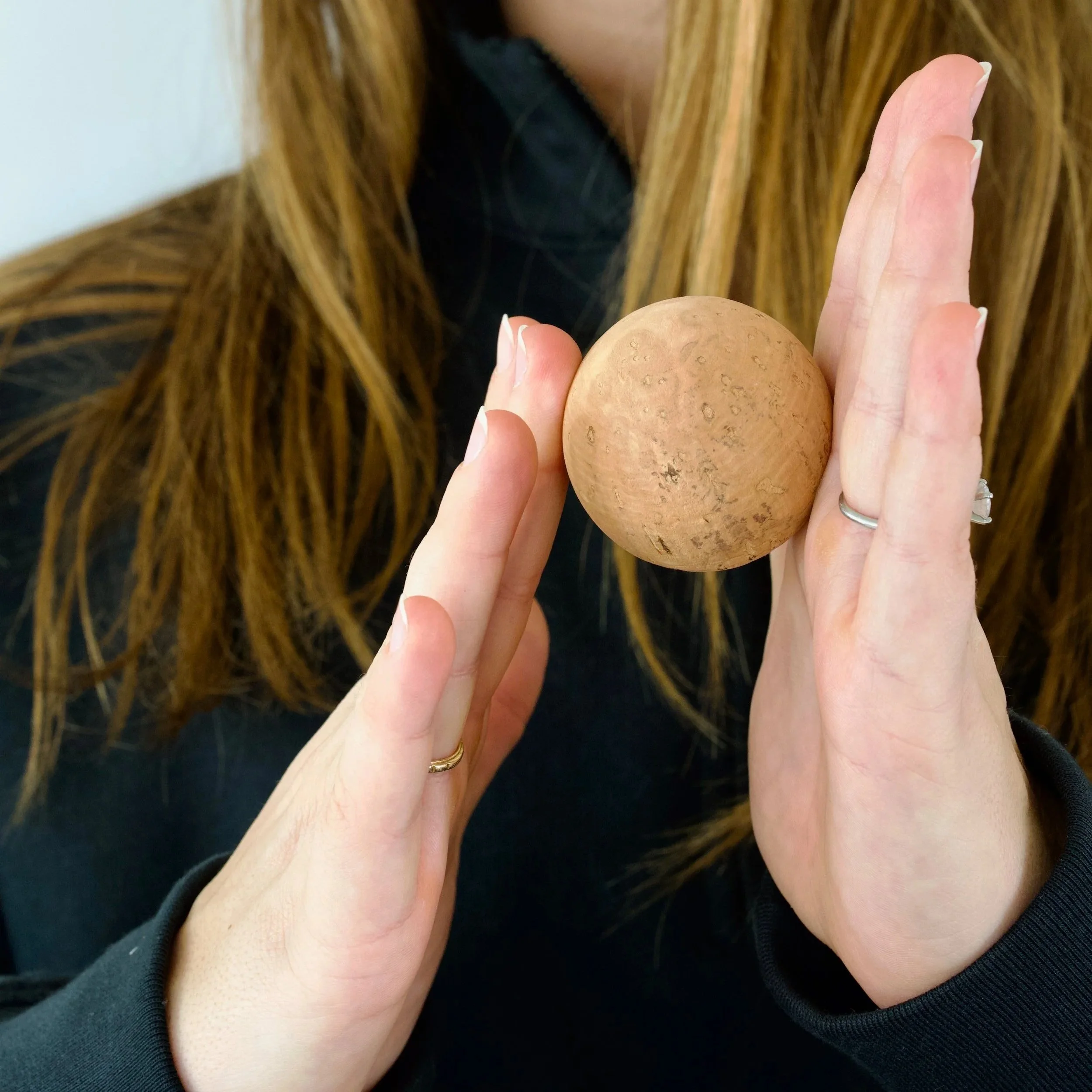 Person holding a small brown spherical object with a textured surface, possibly a nut or seed, with both hands, wearing rings, with long brown hair and dark clothing.