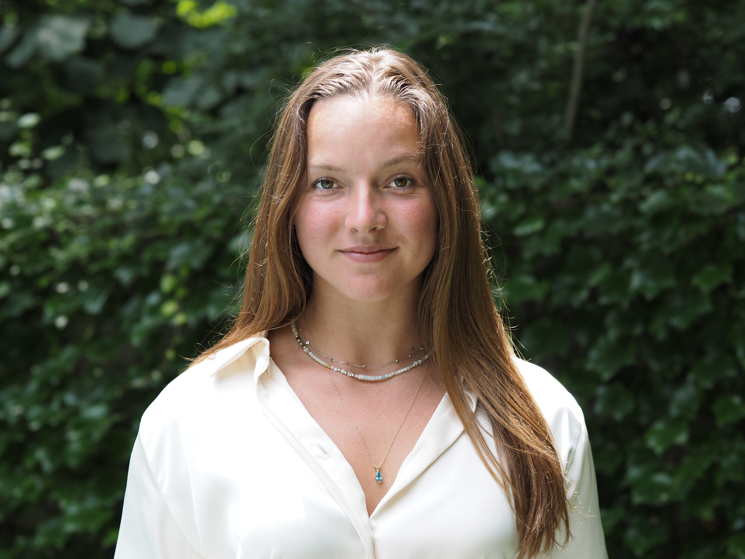 A woman with long brown hair wearing a white blouse and layered necklaces standing in front of green leafy bushes.