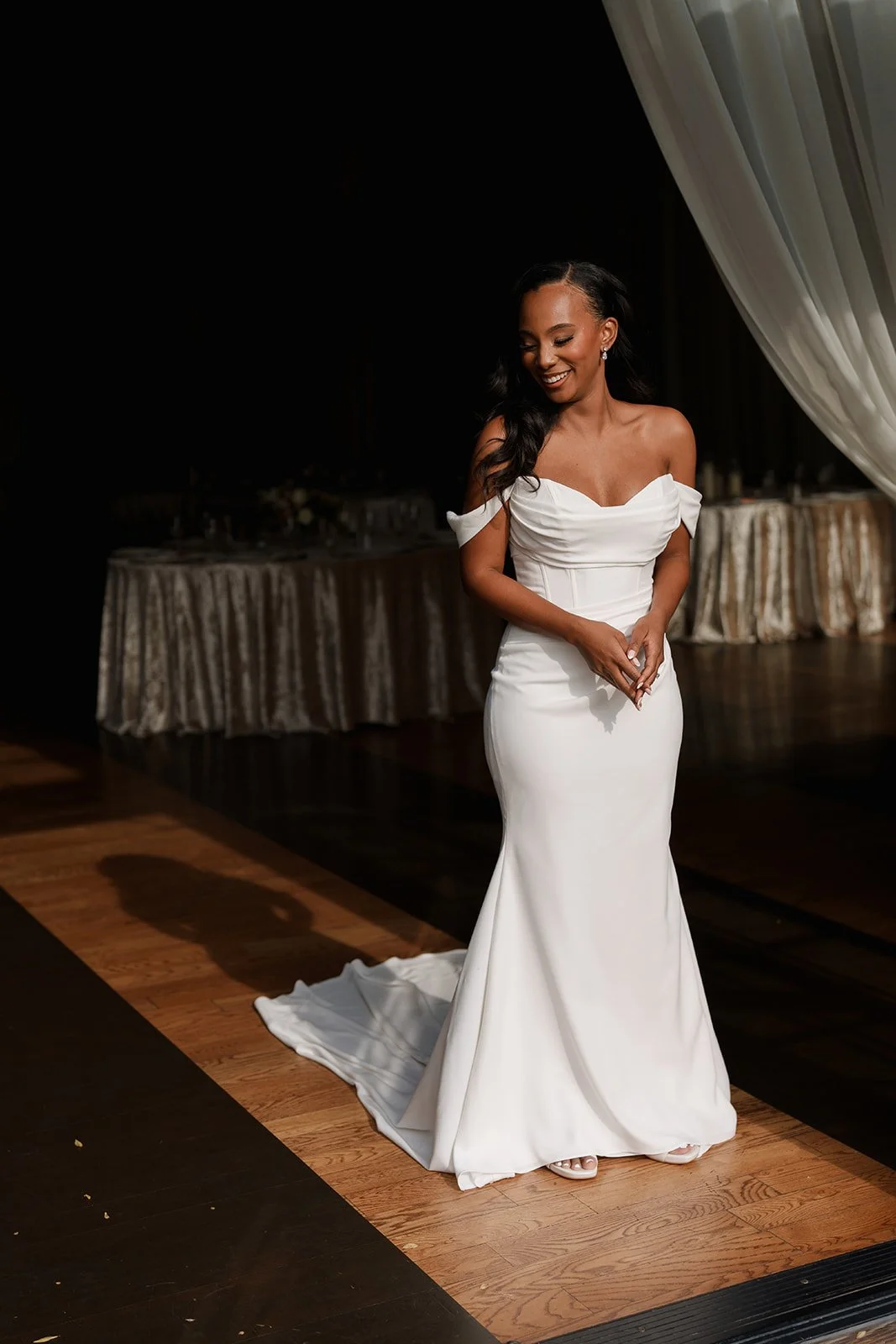 A bride wearing a white off-shoulder wedding gown standing on a wooden floor in a dimly lit room.
