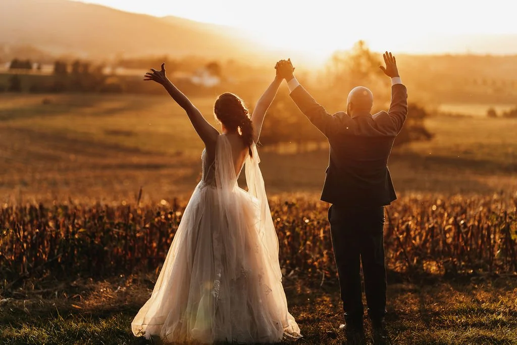 A couple dressed in wedding attire standing in a field at sunset, raising their arms in celebration.