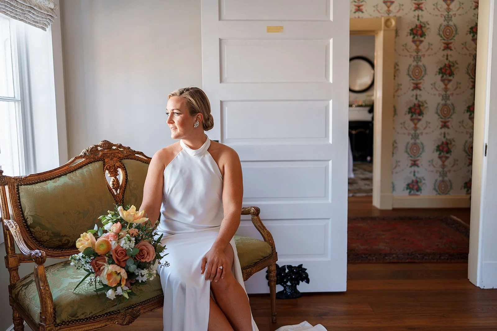 A woman in a white dress sitting on a vintage green upholstered sofa with a bouquet of pink and cream flowers, looking out a window.