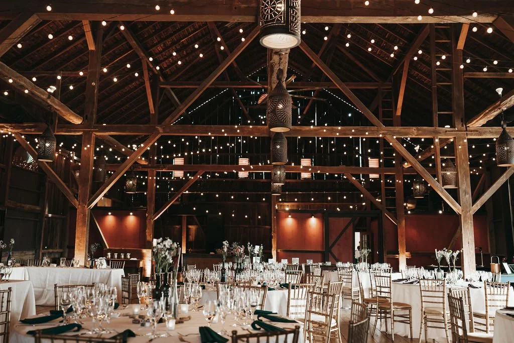 Interior of a rustic event hall with wooden beams, string lights, and tables set for a celebration, with chairs, glassware, and floral centerpieces.