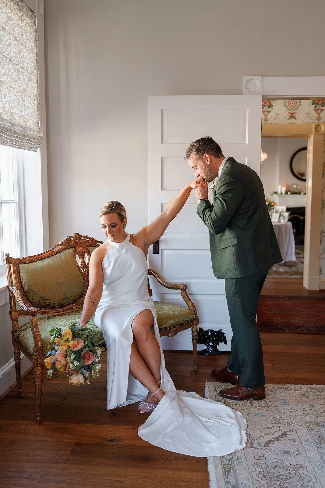 A man in a green suit kisses the hand of a woman in a white dress sitting on an ornate wooden bench, holding a colorful bouquet of flowers.