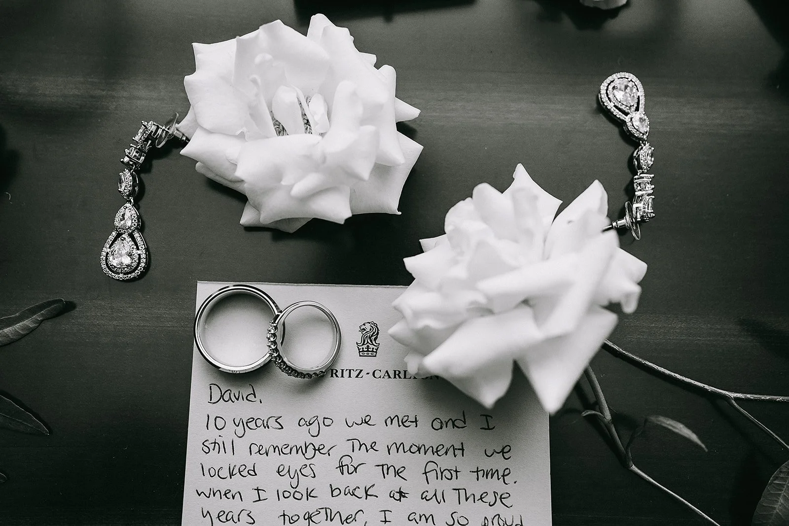 A black and white photo of wedding rings, jewelry, white roses, and a handwritten note on a dark surface.