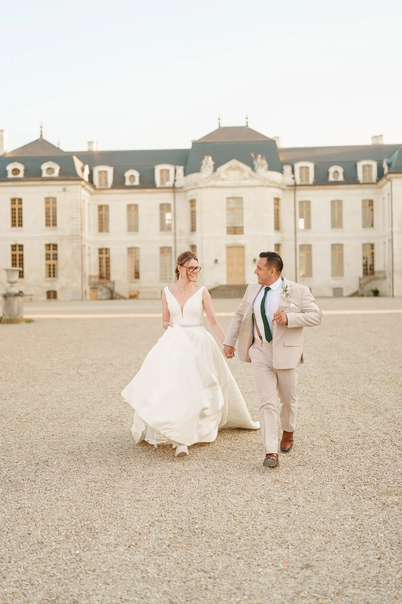 A bride and groom walk hand in hand in front of a historic building, smiling at each other on their wedding day.