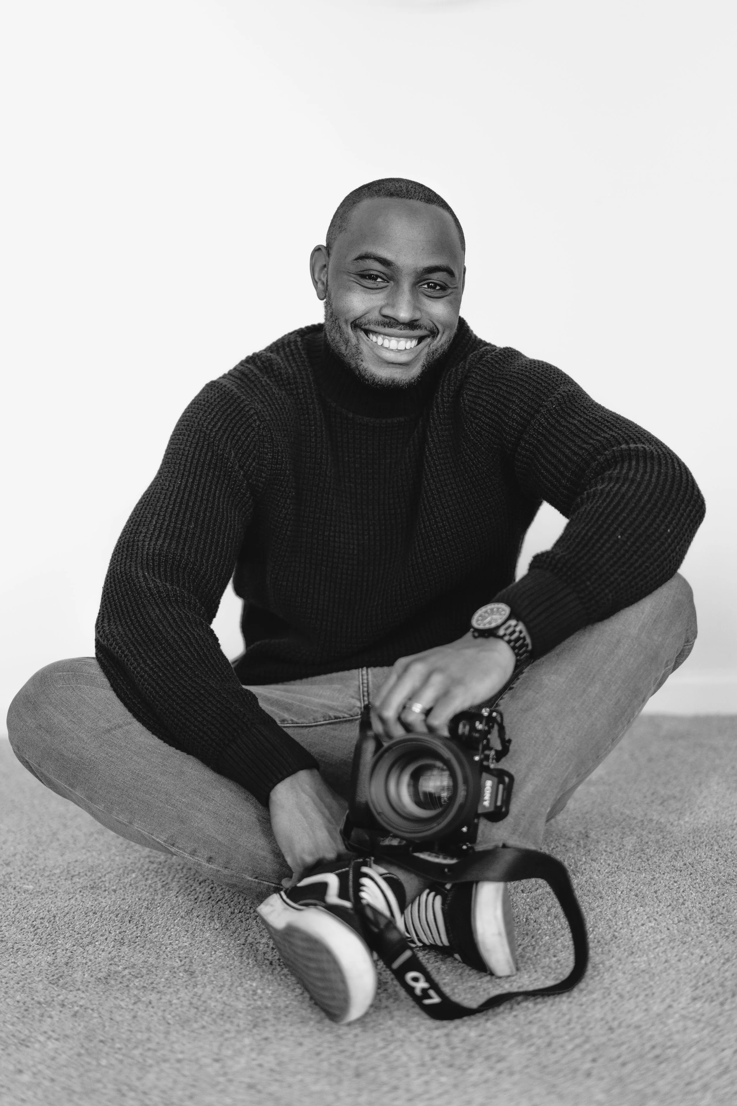 A smiling man in a black sweater and jeans sitting on the floor with a camera in his lap, black and white photo.