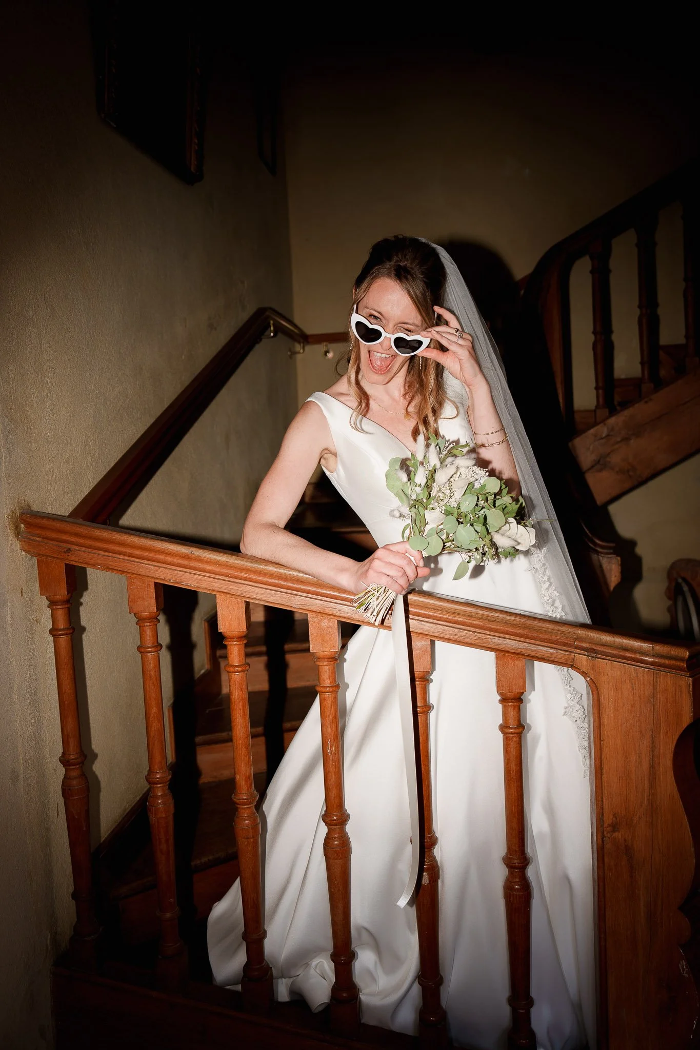 Bride in a white wedding gown holding a bouquet, wearing fun heart-shaped sunglasses, standing on a staircase with wooden banister, smiling and enjoying her special day.