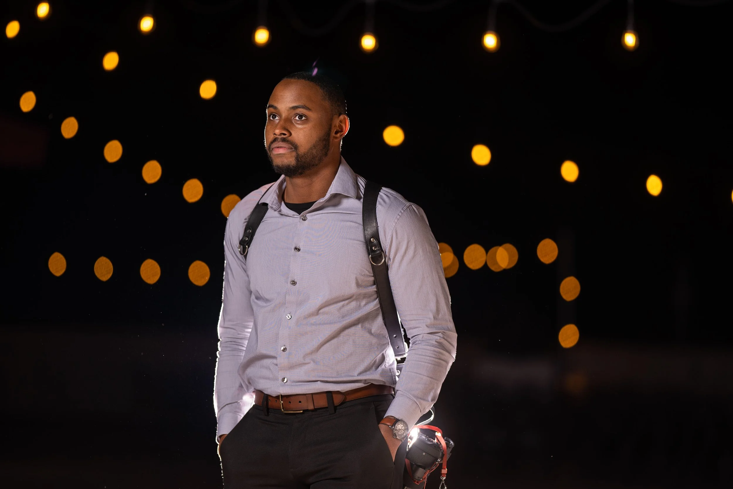 A young man with a beard and mustache, wearing a light purple button-up shirt and black pants, stands outdoors at night with a camera in his right hand and a black backpack over his shoulders. The background features warm yellow bokeh lights.