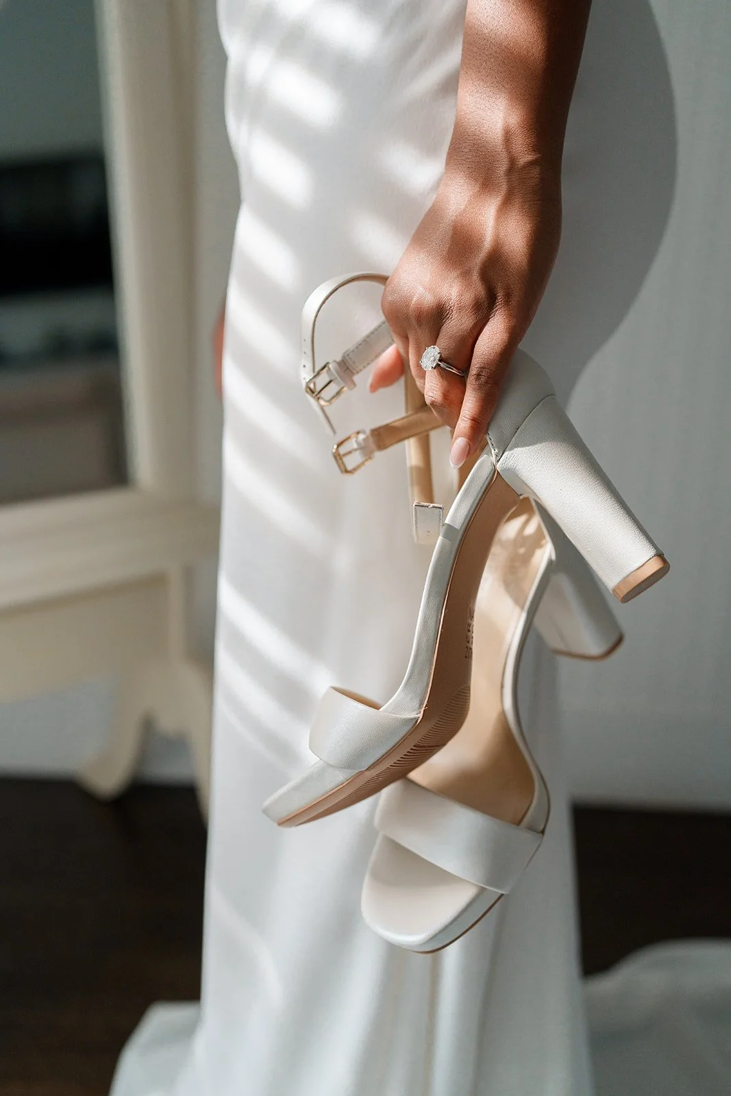 A woman in a white dress holds a pair of white high-heeled sandals with thick straps and a block heel, with sunlight and shadows cast on her arm and dress.