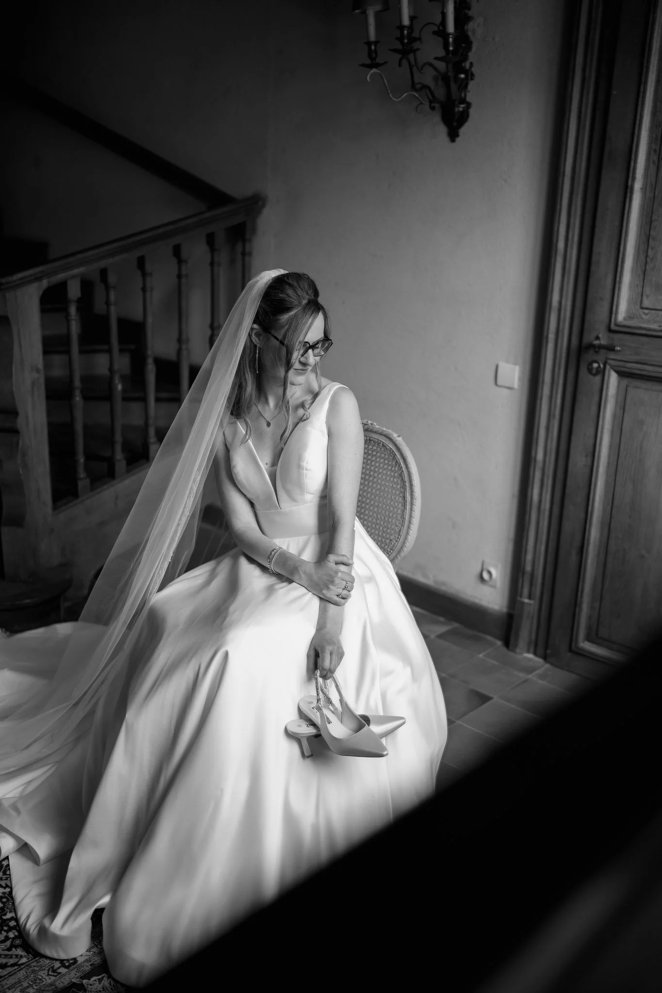 A woman in a wedding dress with glasses, a veil, and jewelry sitting on a chair in a room, holding a pair of high-heeled shoes.
