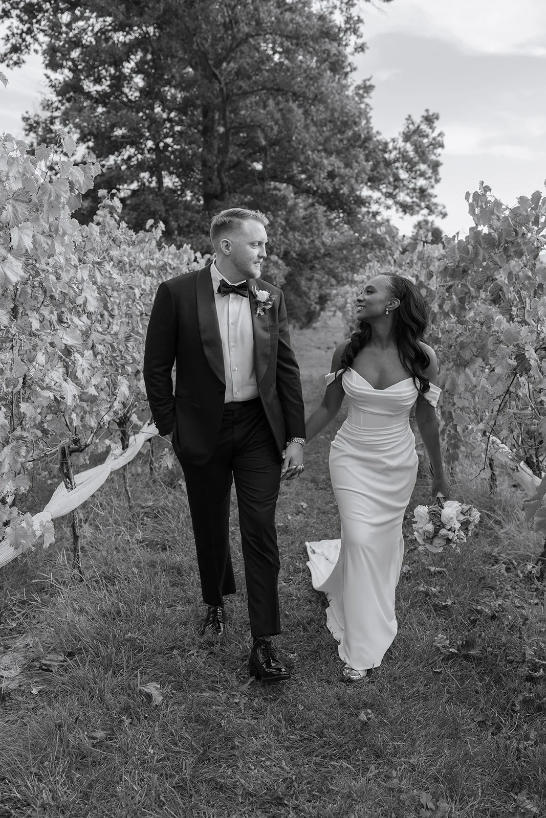 Black and white photo of a bride and groom walking hand in hand through a vineyard. The groom is wearing a tuxedo with a bow tie, and the bride is in a wedding dress holding a bouquet of flowers, both smiling at each other.