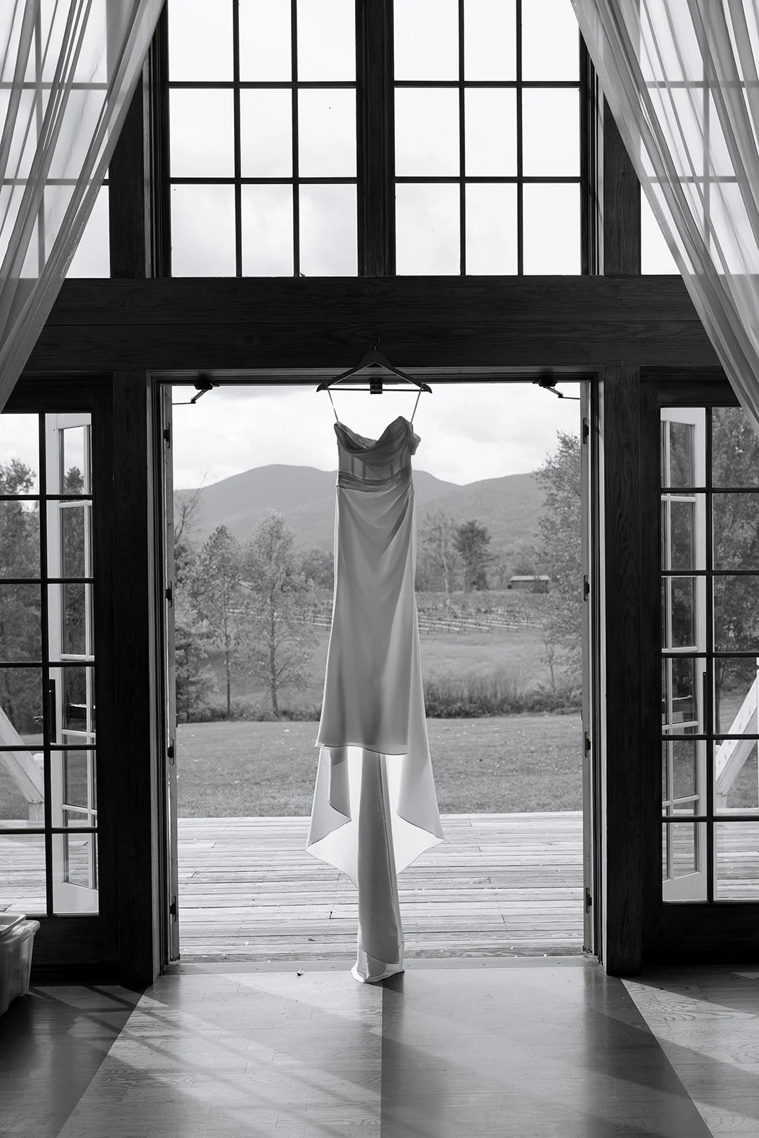 A wedding dress hanging on a hanger in front of open glass doors with a scenic mountain and field view in the background, black and white photo.