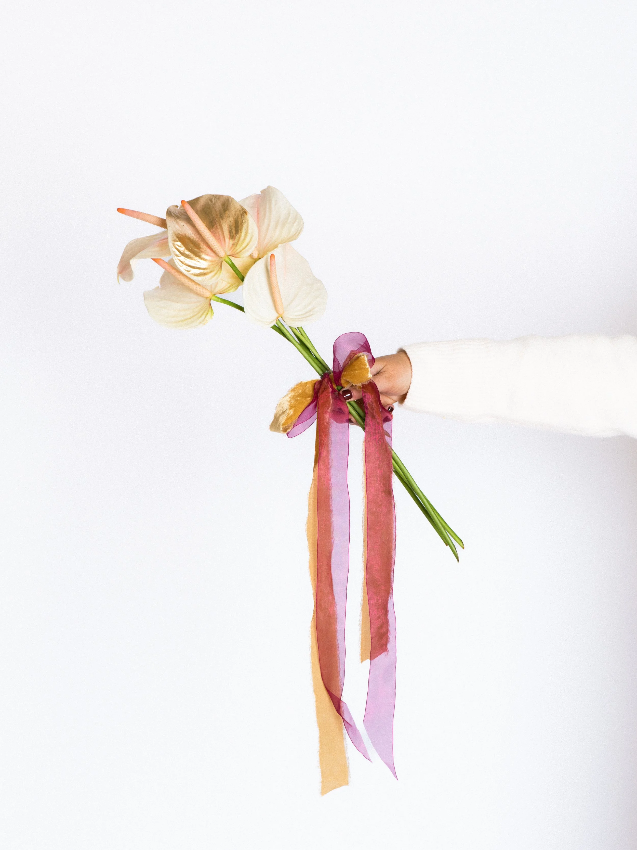 Person holding a bouquet of beige and white flowers tied with pink, purple, and gold ribbons against a plain white background.