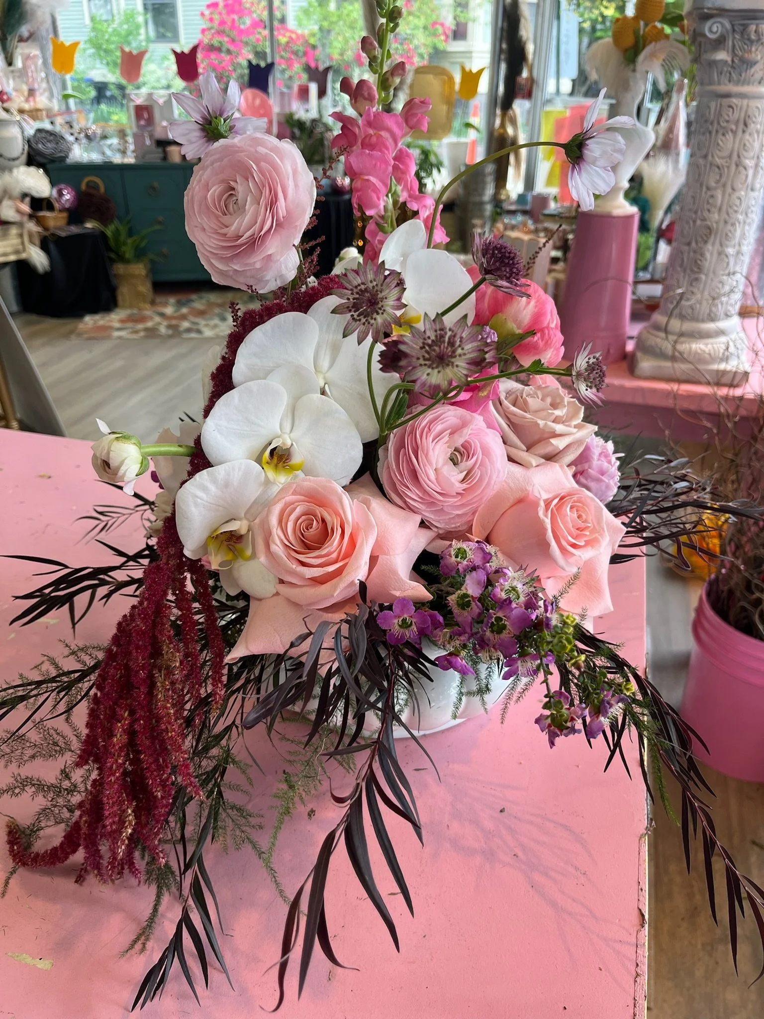 A floral arrangement in pastel pink, white, and purple flowers, including roses, orchids, and other blooms, displayed on a pink table inside a decorated shop with colorful decorations and a window showing a garden outside.