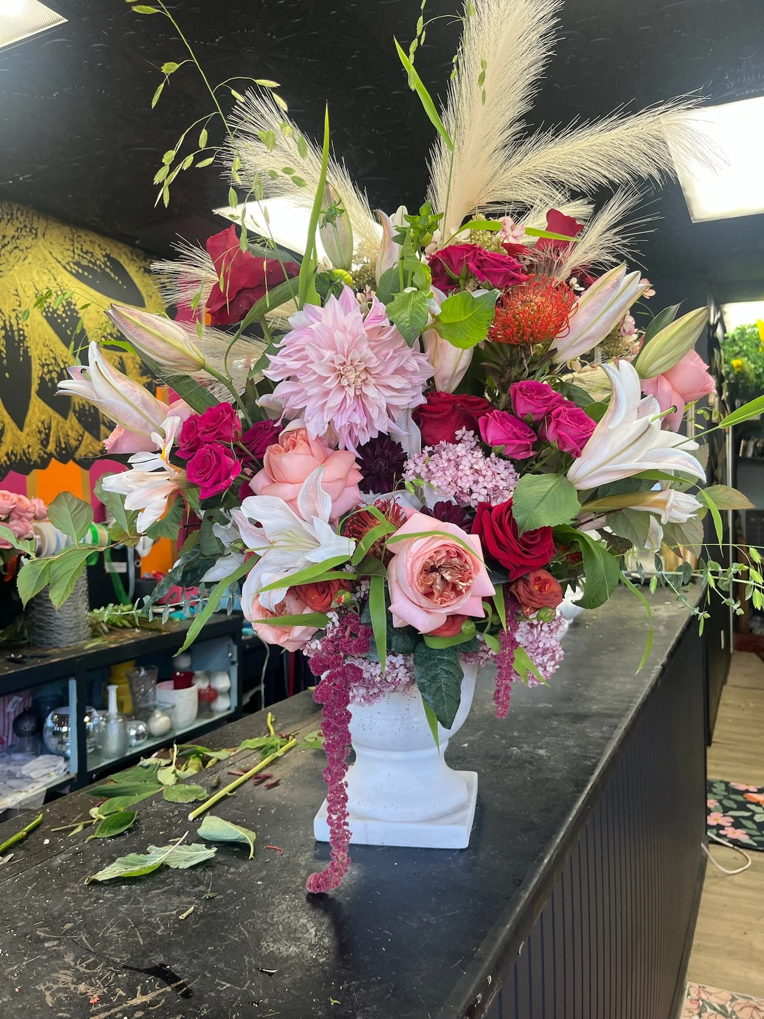 A large, colorful floral arrangement in a white vase with pink, red, and purple flowers, including roses, lilies, and dahlias, along with pampas grass and greenery, placed on a black counter.