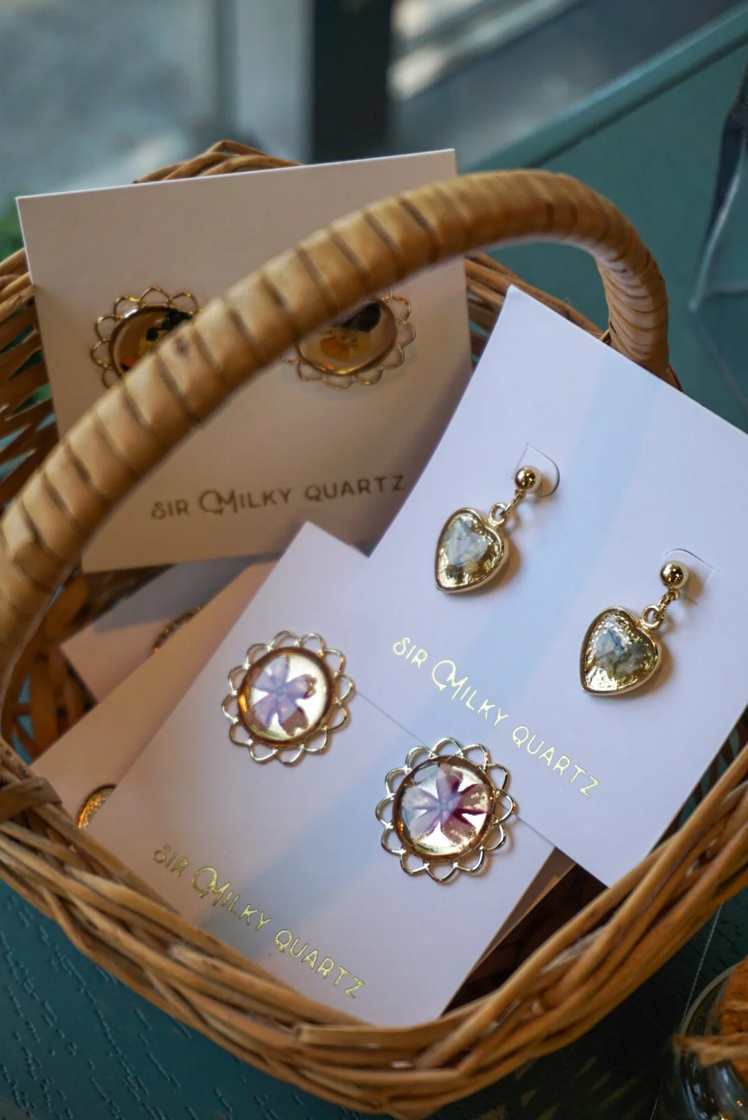 A wicker basket containing earrings from Sir Milky Quartz, including heart-shaped and floral designs, displayed on white cards with gold lettering.